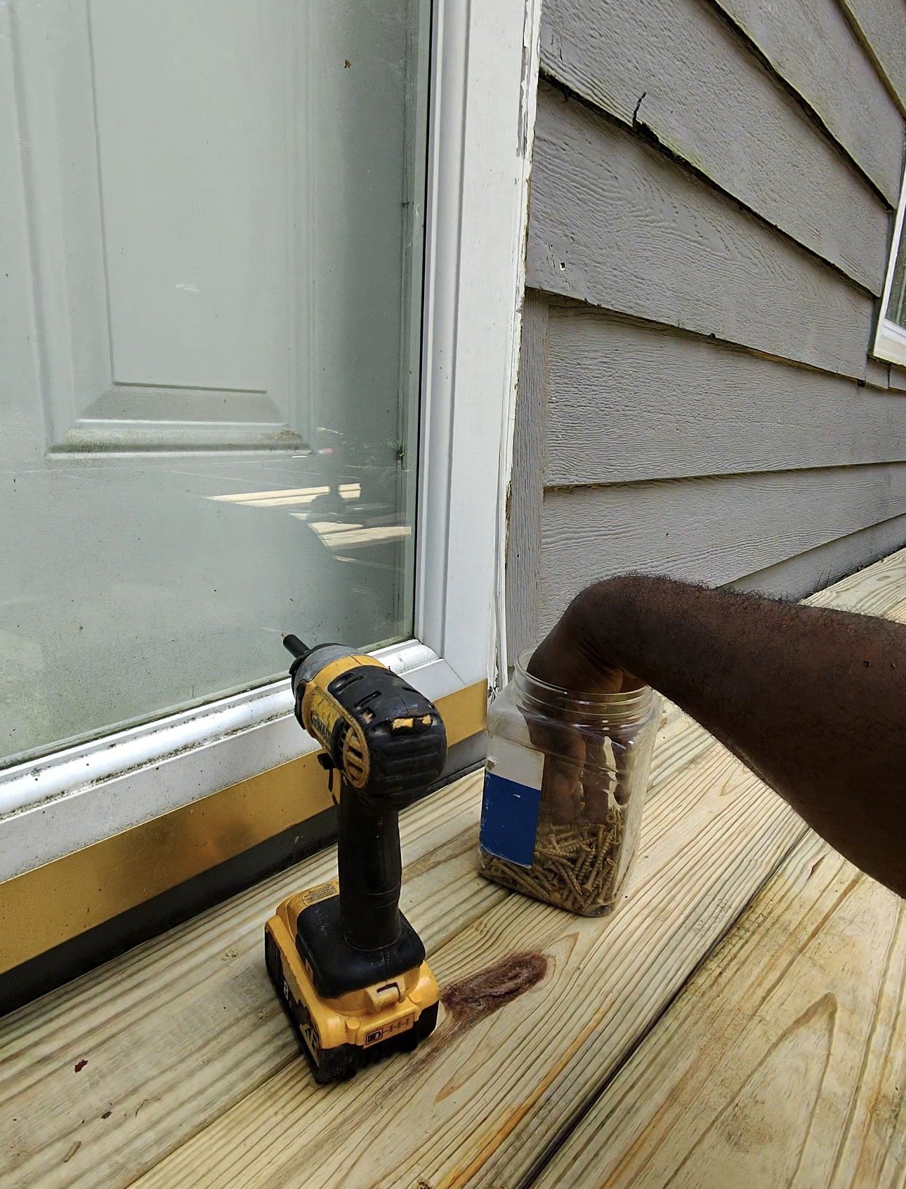 A person's arm is visible as they use a cordless drill near a door frame on a wooden deck. There is a container of screws and a cordless drill on the deck, along with a window and house siding in the background.