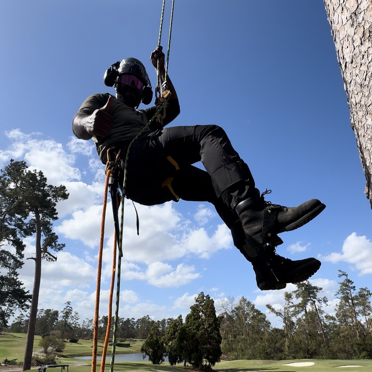 A person wearing a helmet, safety goggles, and harness is rappelling or performing tree work outdoors during the daytime, with a clear blue sky and trees in the background.
