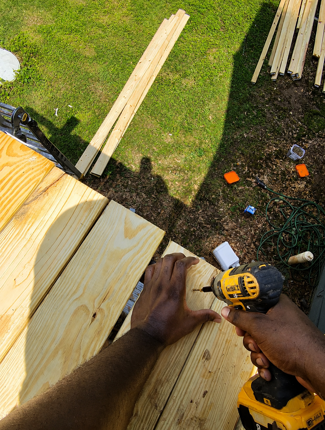 Person using a cordless drill to screw a piece of wood onto a wooden structure outdoors with grass and scattered tools around.