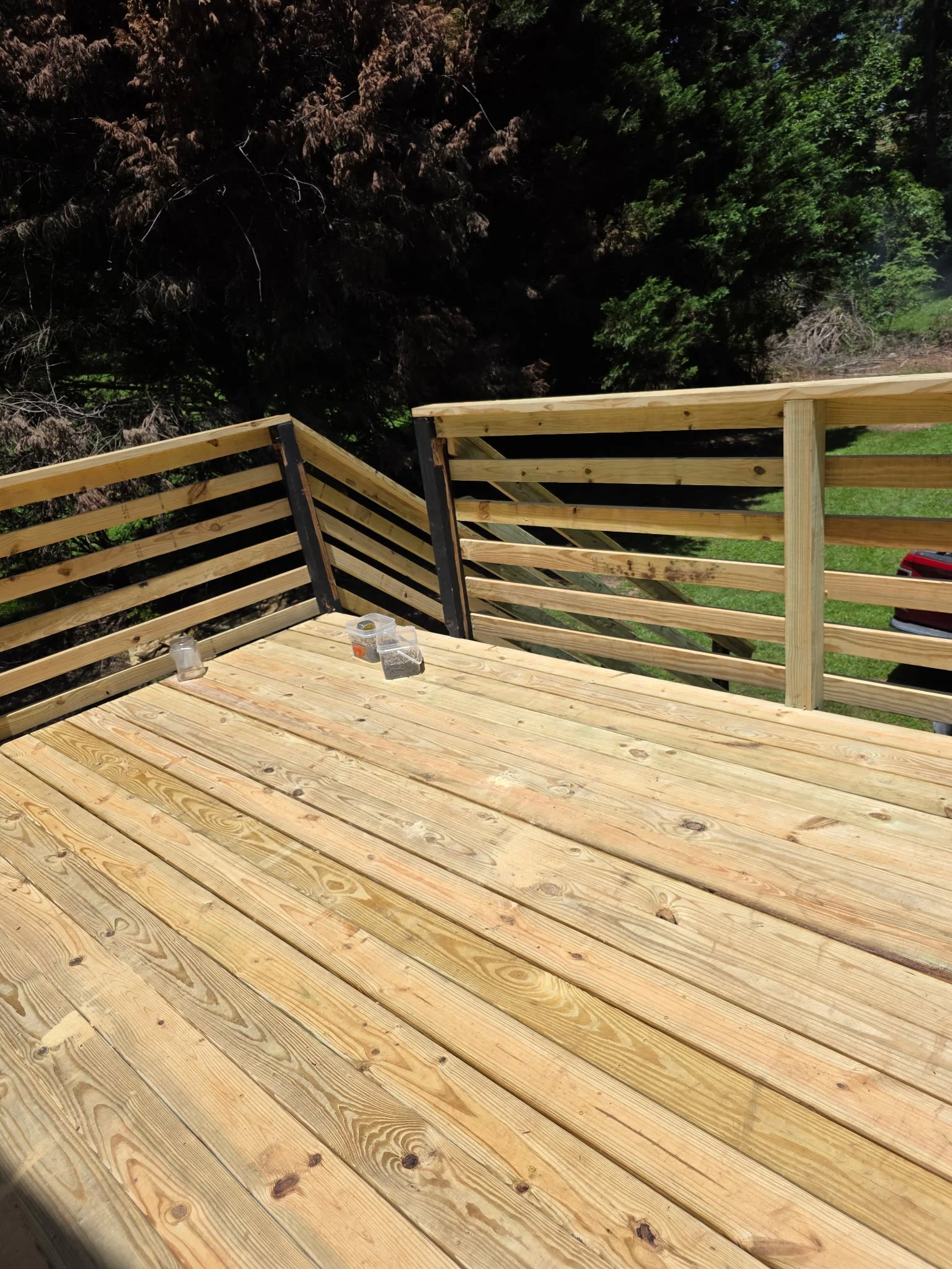 Wooden deck construction with partially installed railing, tools and screws on the deck, green trees and grass in the background.