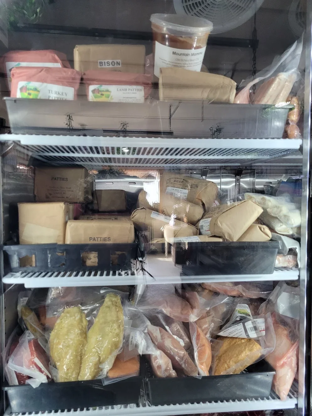 Frozen meats and food items in a glass-front refrigerator, including packages labeled patties, bison, turkey, lamb, and various other meats, some in plastic wrap and boxes.