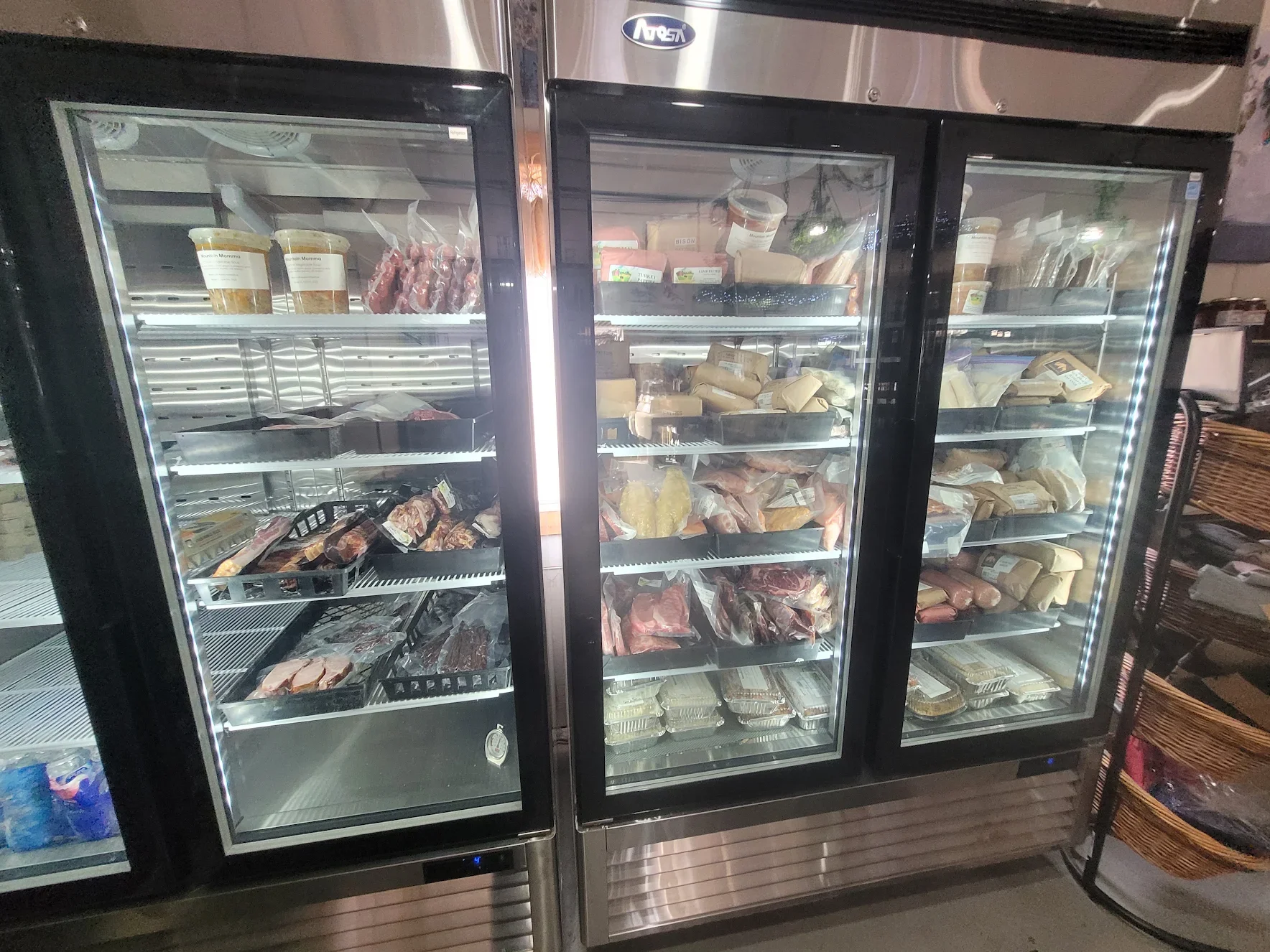 A large commercial refrigerator with glass doors filled with various frozen meats and packaged food items. The refrigerator is located in a grocery store or similar retail environment.