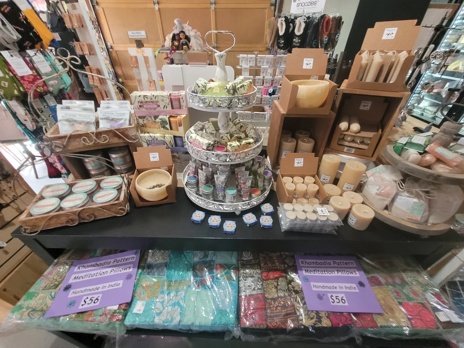 Display of handmade meditation pillows, candles, and fabric patterns in a store. Items include fabric-covered pillows, candles, and handmade accessories, with price tags indicating $56.