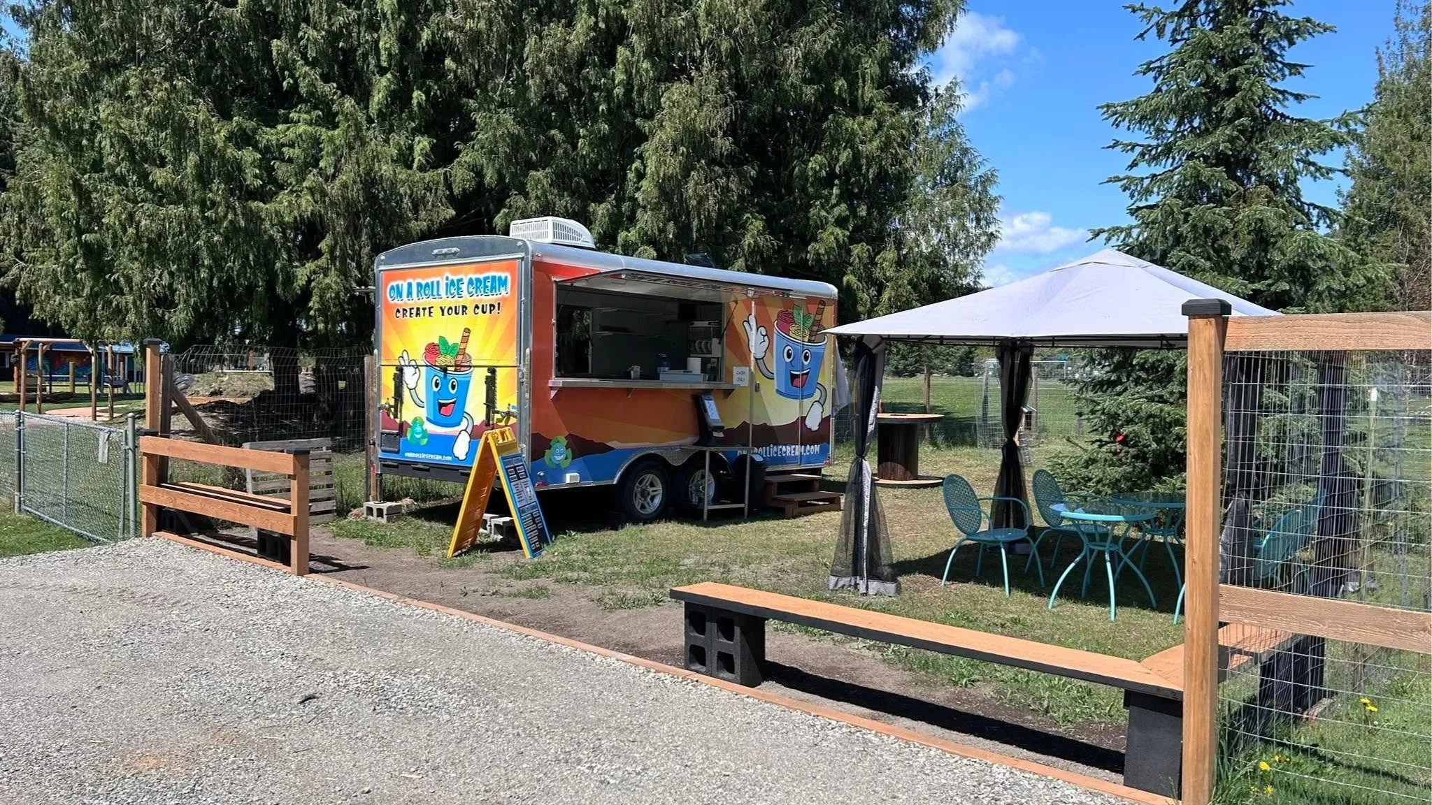 A colorful ice cream truck labeled "On a Roll Ice Cream" with a character holding a cone, parked next to a shaded outdoor seating area with tables, chairs, and a canopy in a park setting with trees and blue sky.