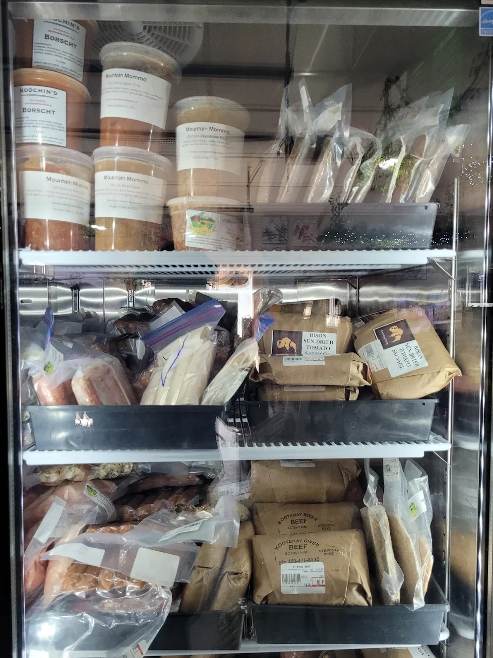 A fridge filled with various packaged foods, including containers of Mountain Momma chili, vacuum-sealed meats, and paper-wrapped products, with some products labeled as beef and other ingredients.