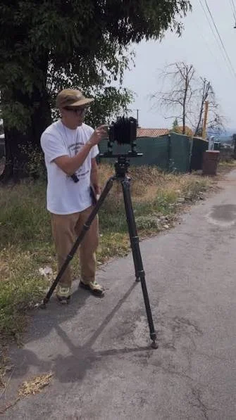 Person operating a camera on a tripod outdoors on a street with trees and power lines.