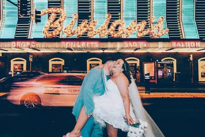 Couple sharing a kiss outside a neon-lit building with signage that reads "Budds" and other illuminated signs, with a taxi cab in the foreground.
