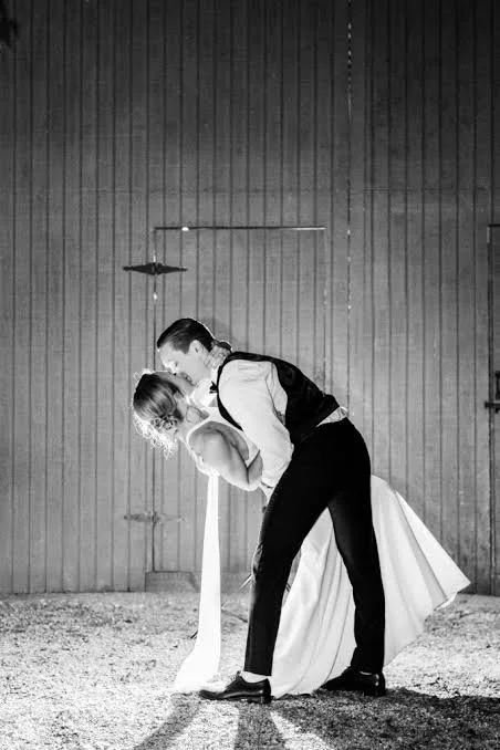 A black and white photo of a couple dancing closely, with the man dipping the woman backward, against a wooden wall background.