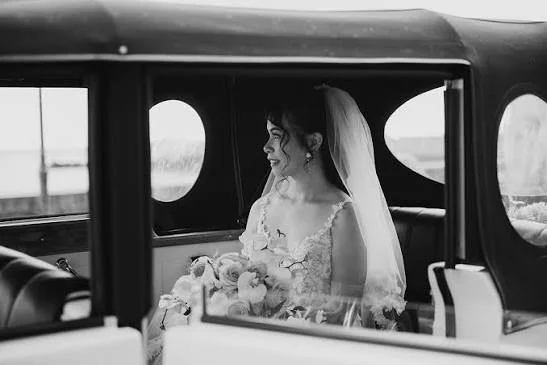 A bride sitting inside a classic car, wearing a wedding dress and holding a bouquet.