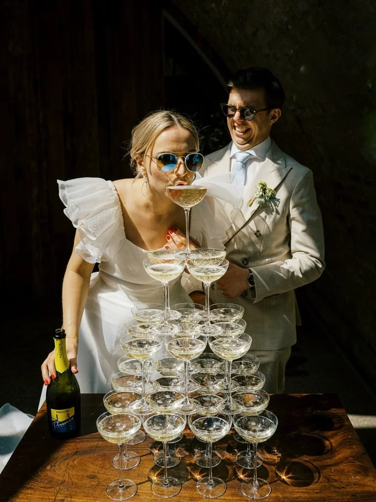 A couple dressed in wedding attire standing behind a pyramid of champagne glasses, with the woman about to drink from the top glass, and the man smiling and wearing sunglasses and a boutonniere.