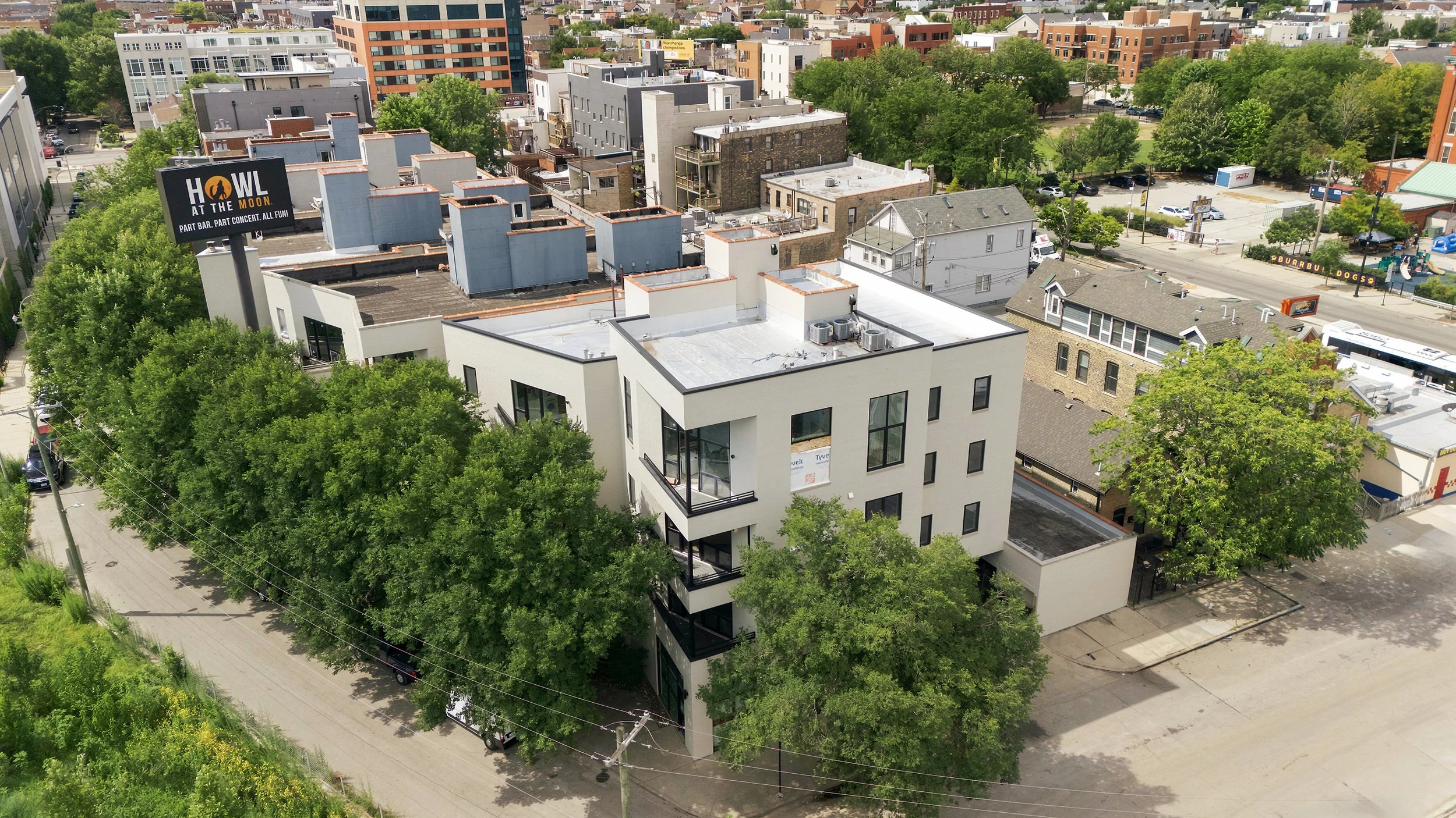 Modern 4-story apartment building with dark brick exterior, large windows, and small balconies at each level, situated on a city street with trees and cars.