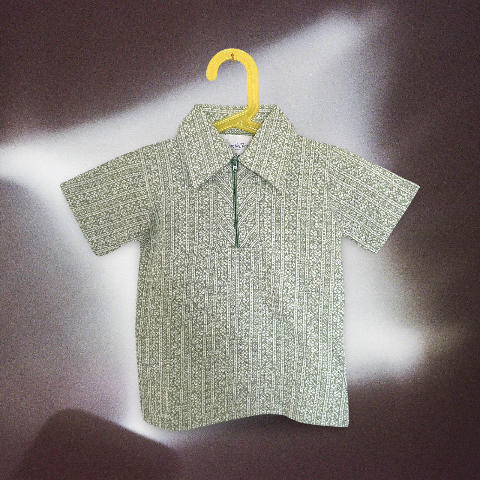 Vintage 1970s green and white patterned short-sleeved shirt with a zipper on the front, hanging on a yellow hanger against a dark and light background.