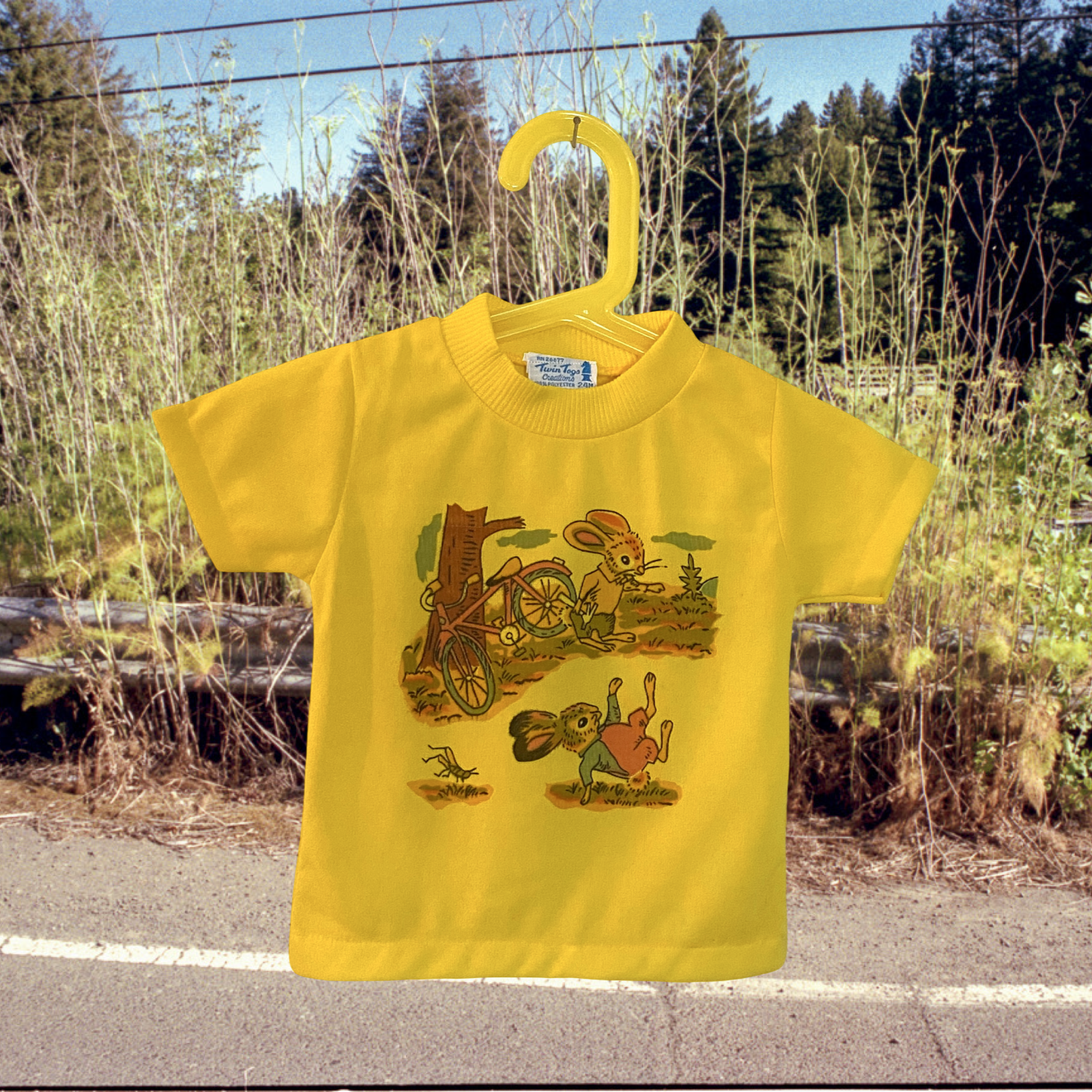 Vintage 1960s Yellow T-shirt featuring a cartoon illustration of a rabbit and a mouse riding a bicycle, hanging on a yellow hanger outdoors against a background of trees and power lines.