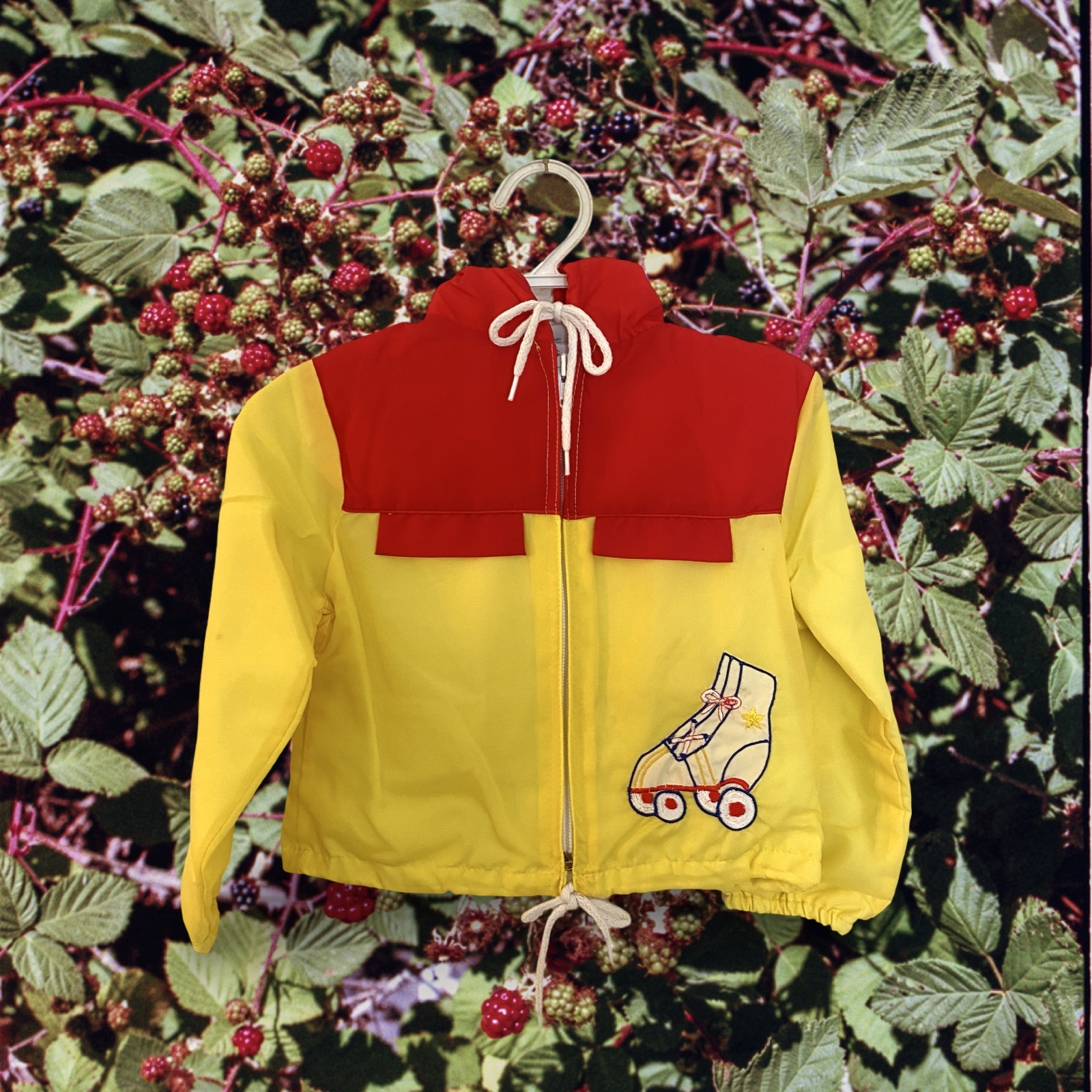 Vintage 1970s bright yellow jacket with a red yoke and a white drawstring hood hanging on a white hanger in front of a background of blackberry bushes.