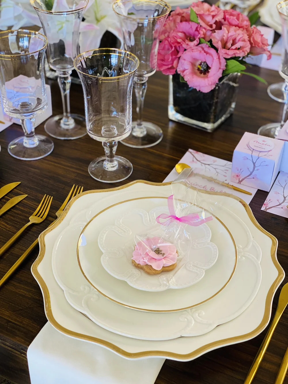 Puesto de mesa con vajilla blanca y dorada, una flor rosa y tarjeta con lazo junto a un ramo de peonías