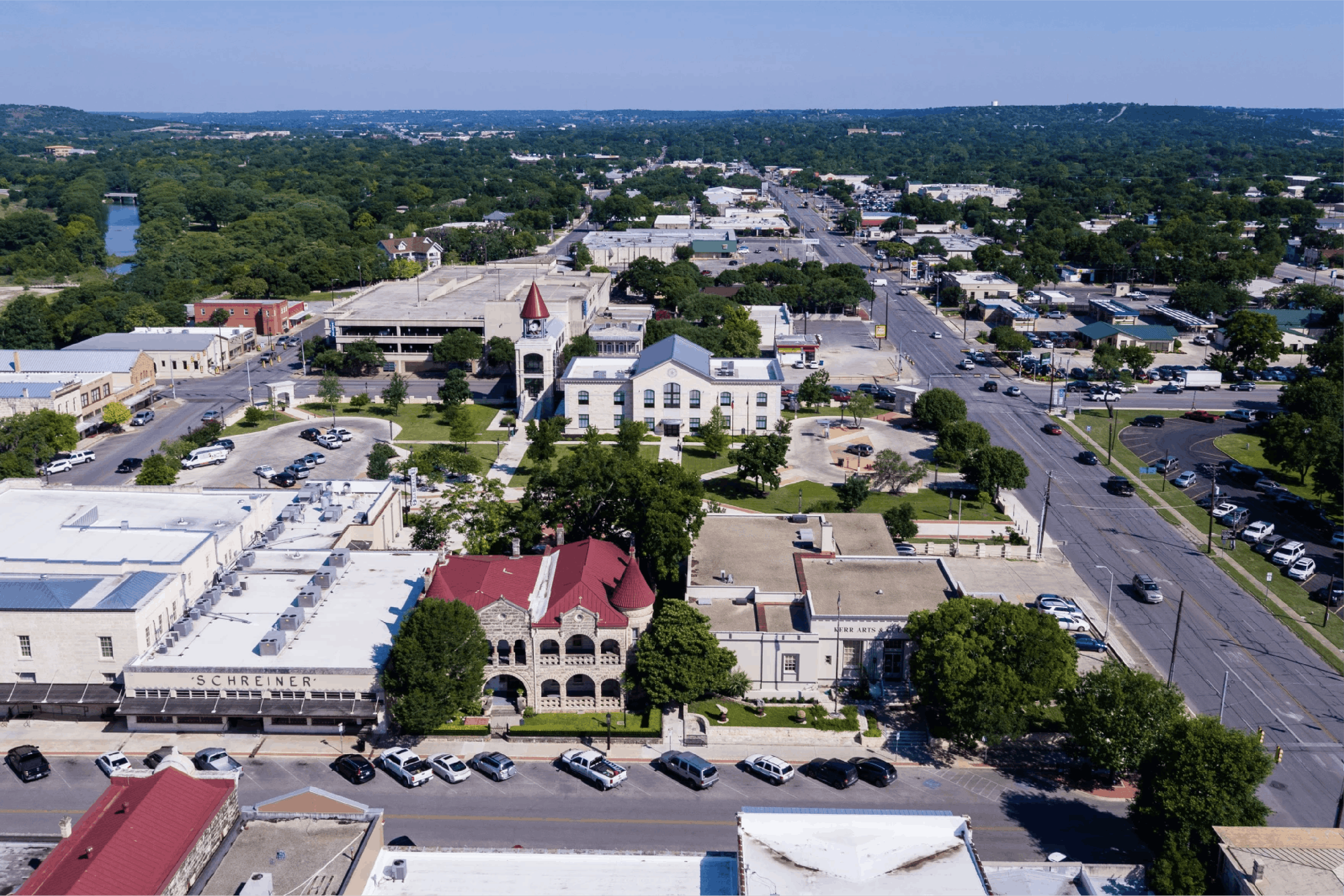 An aerial view of a Riverfront Village with buildings, streets, parking lots, trees, and a river surrounded by greenery, under a clear blue sky.