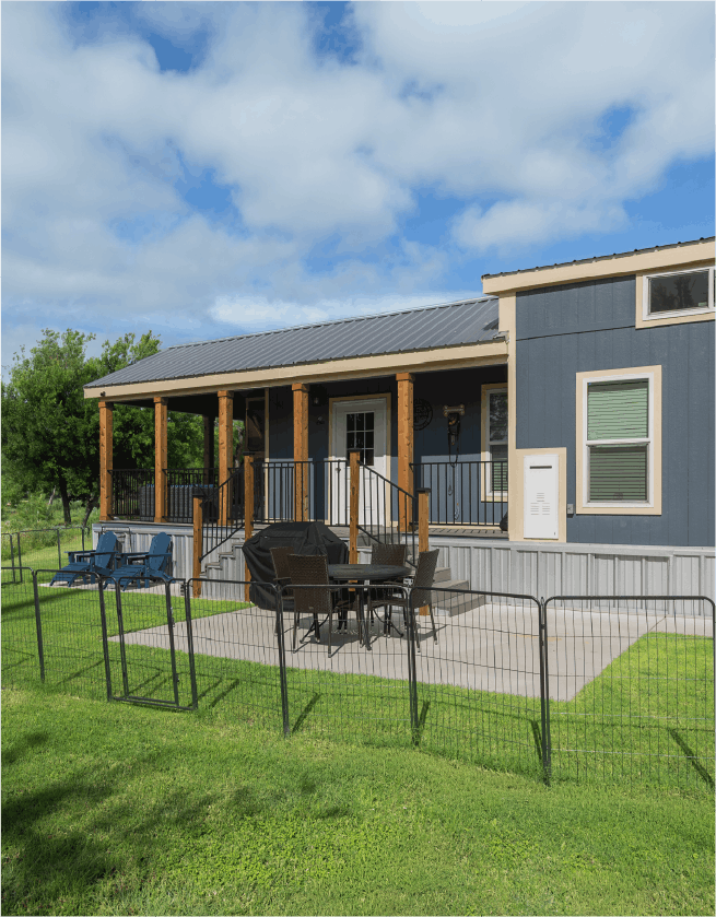 Backyard patio with outdoor furniture, a grill covered with a black cover, and a small fenced yard in front of a blue house with a porch, under a partly cloudy sky.