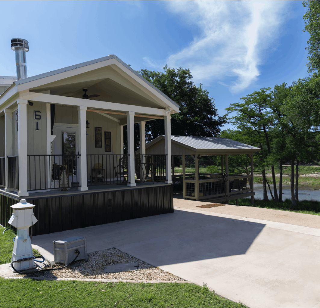 A tiny home with a covered porch and a smaller outdoor structure next to it, situated near a river with trees in the background, under a bright blue sky with scattered clouds.