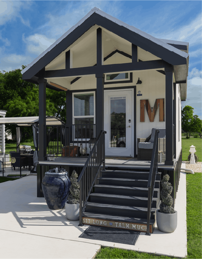 Front porch of a tiny home with black and white exterior, stairs, potted plants, and outdoor seating at Riverside Village.