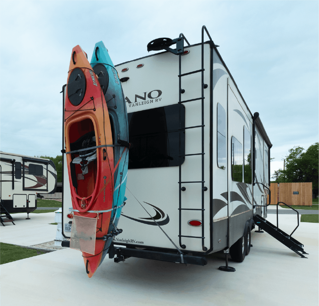Travel trailer with two kayaks mounted on the back, one in orange and the other in blue, parked at a concrete site at Riverfront Village on a cloudy day.