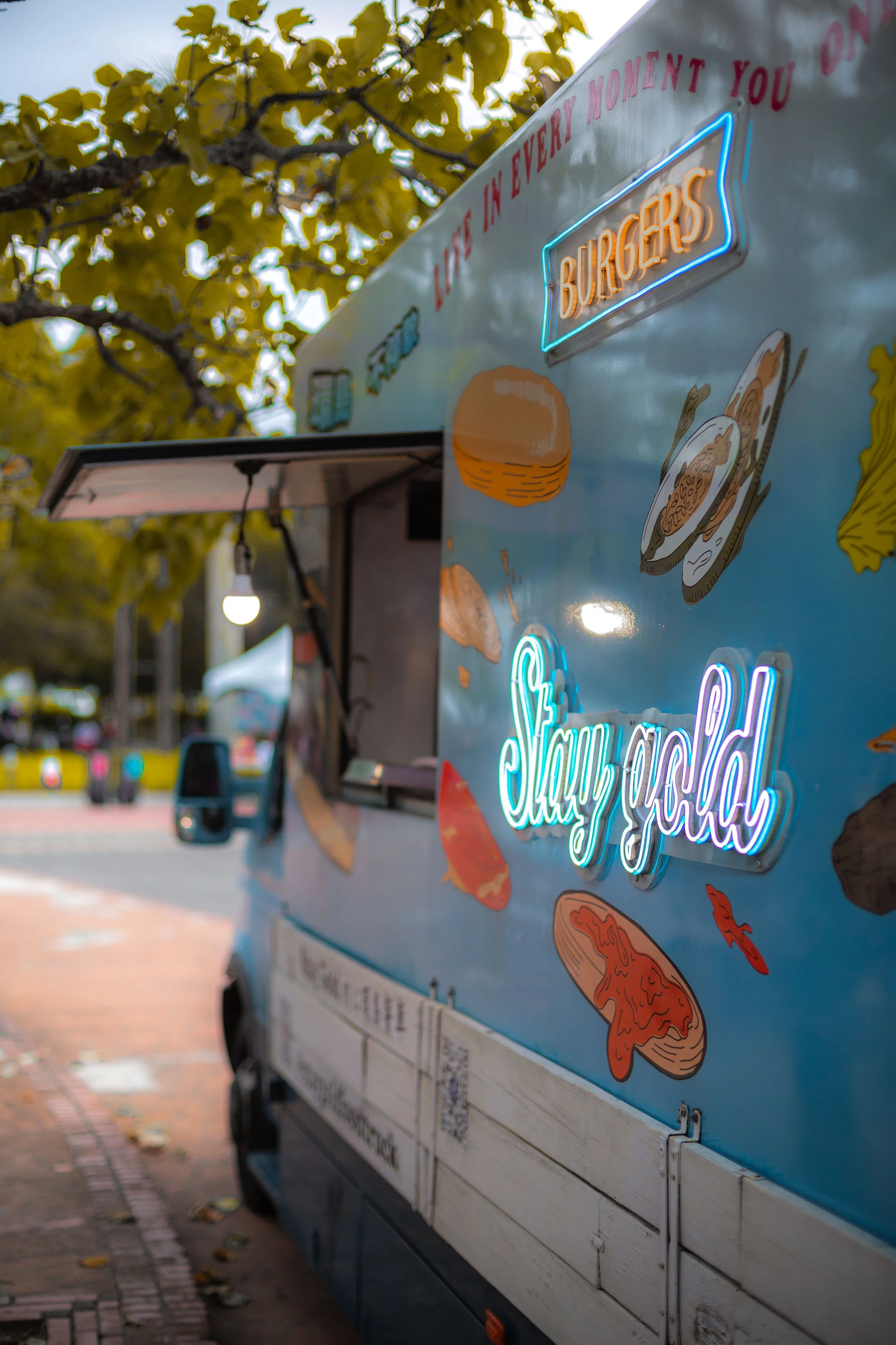 A food truck with a menu and neon sign for burgers, parked outside on a street lined with trees with yellow leaves, during daytime.