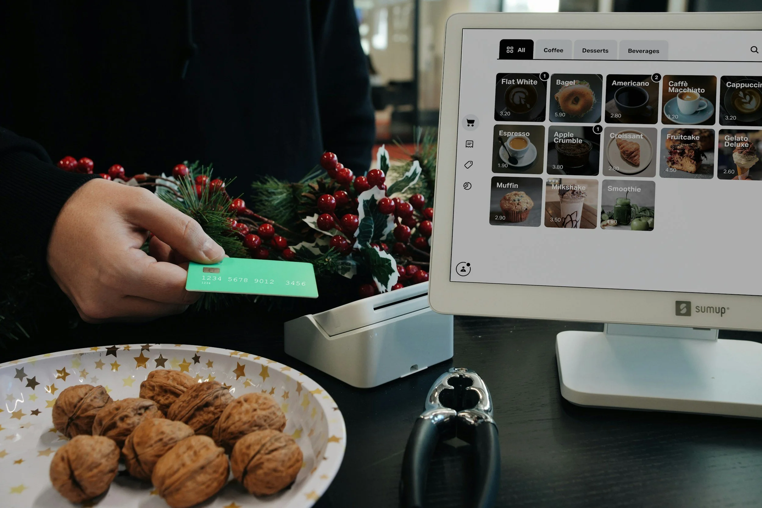 A person holding a credit card in front of a Christmas holiday wreath with red berries and green foliage. A computer screen displays a digital menu of beverages and desserts, including coffee, espresso, croissant, fruitcake, and gelato. On the table, there is a plate of whole walnuts and some cookies.