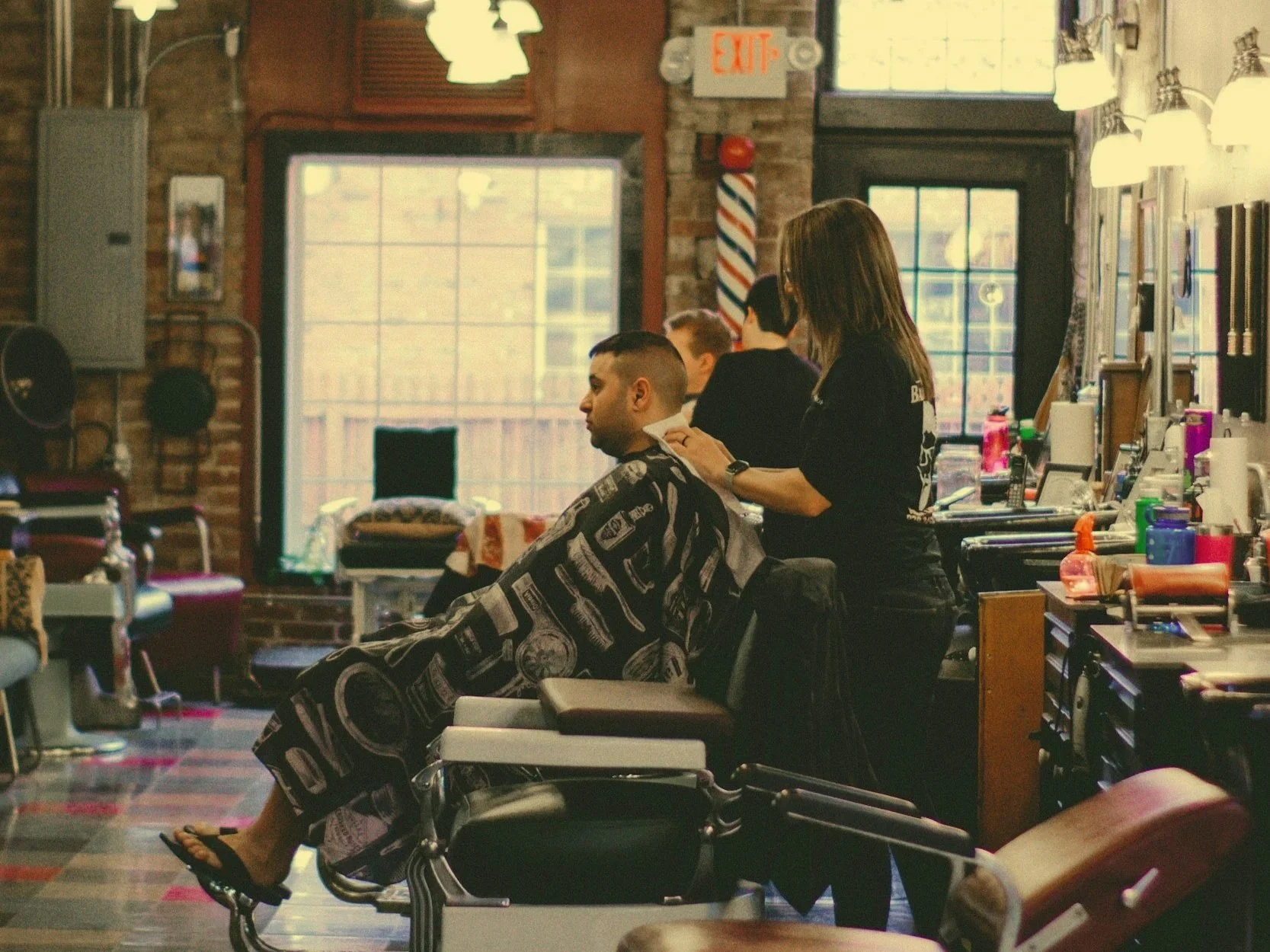 A man getting a haircut in a barbershop, sitting in a barber chair covered with a black and white barber cape, while a female barber styles his hair. The barbershop has wooden accents, large windows, and various barber tools and products on the counters.