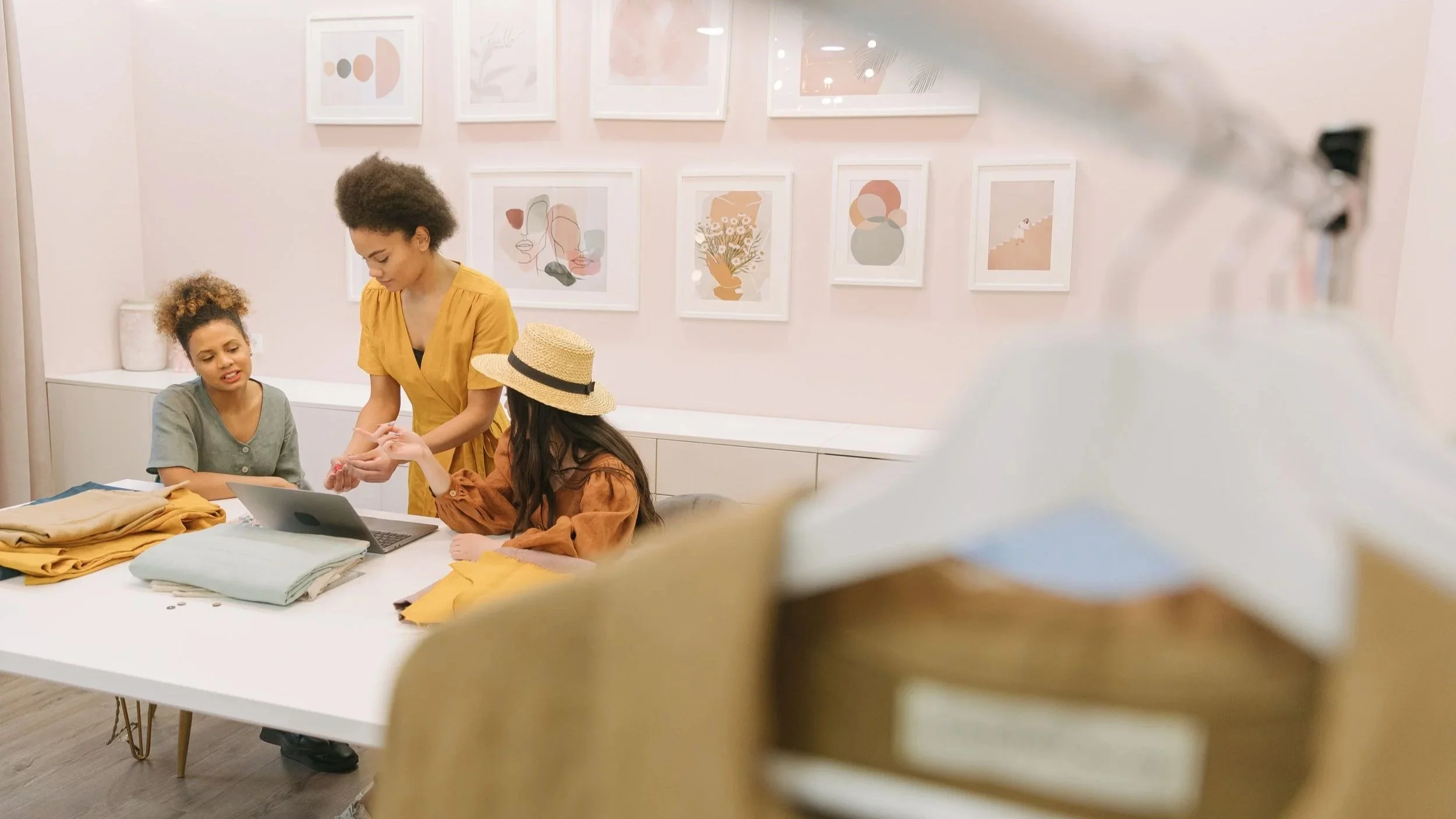 Three women interacting around a table in an art gallery or studio. One woman standing and two women seated, with fabric and a laptop on the table. Artwork is displayed on the wall behind them.