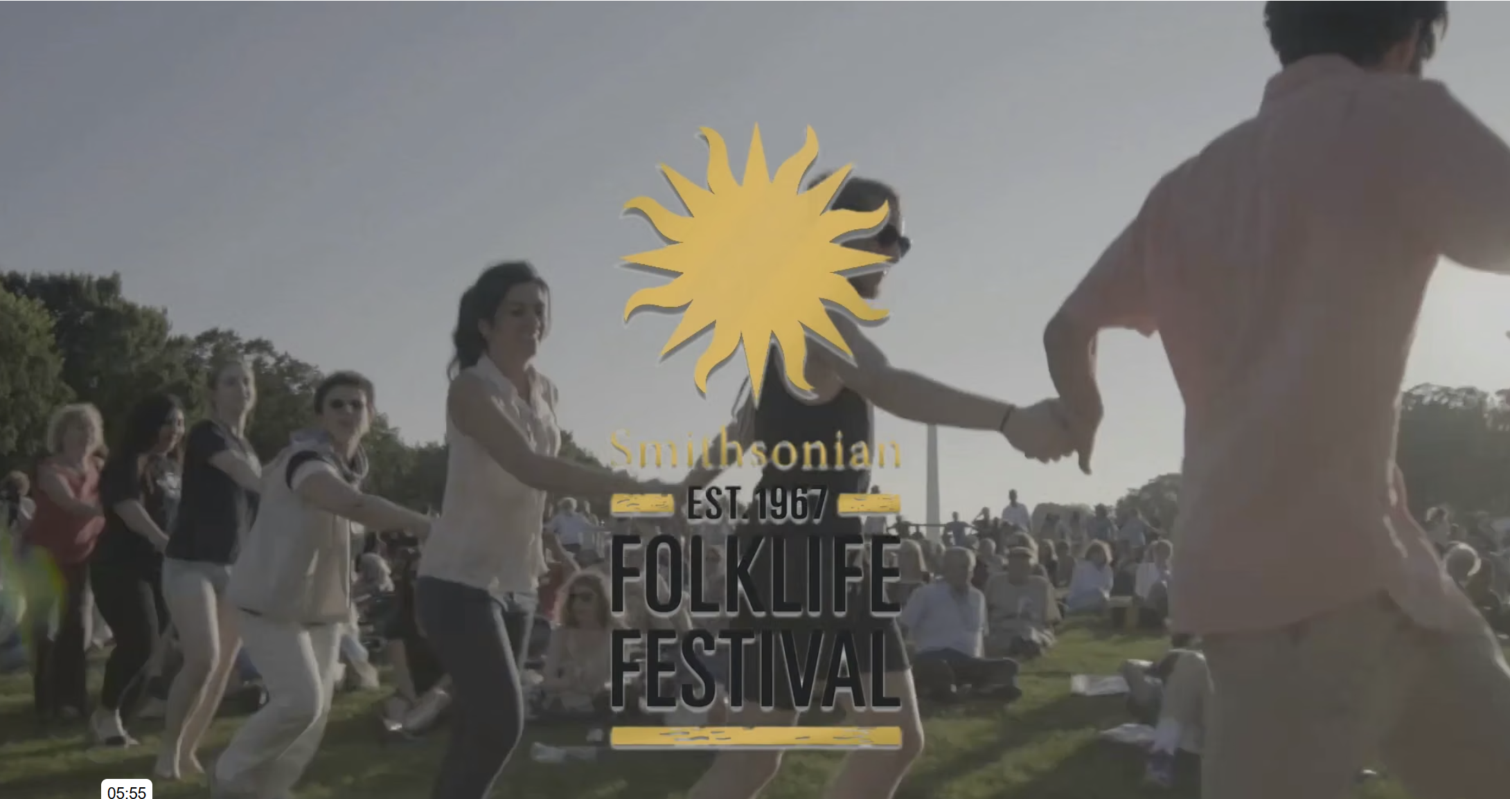 A crowd of people, including several women in skirts and casual wear, gather outdoors on a sunny day. Some people are holding hands, forming a circle, while others stand or sit around, watching the event. The background shows trees and a clear sky. A large overlay features a yellow sun emblem with the text 'Smithsonian Est. 1967 Folklife Festival'.