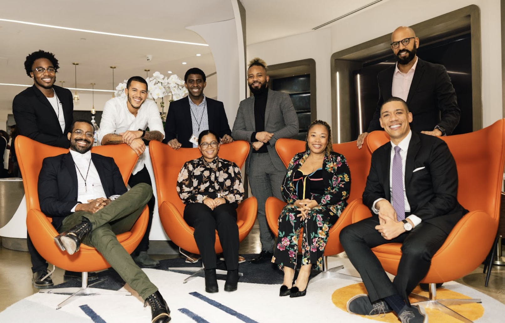 Group of ten diverse professionals posing in a modern office or hotel lobby. Four are seated in orange chairs, and six are standing behind them.