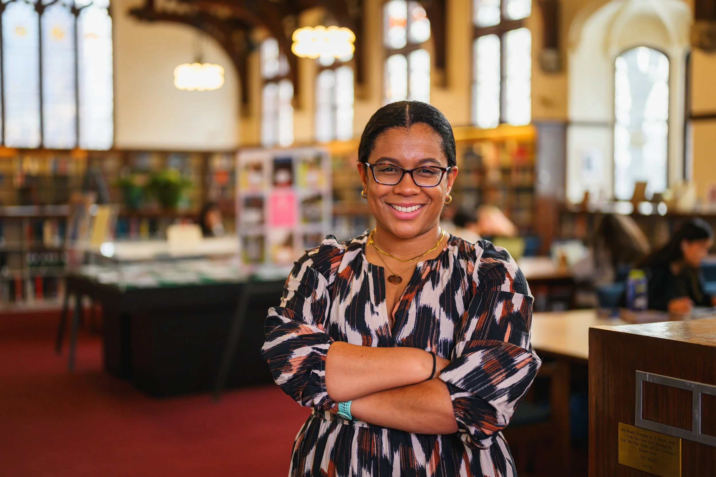 Woman with glasses smiling at the camera inside a library with bookshelves and large windows.