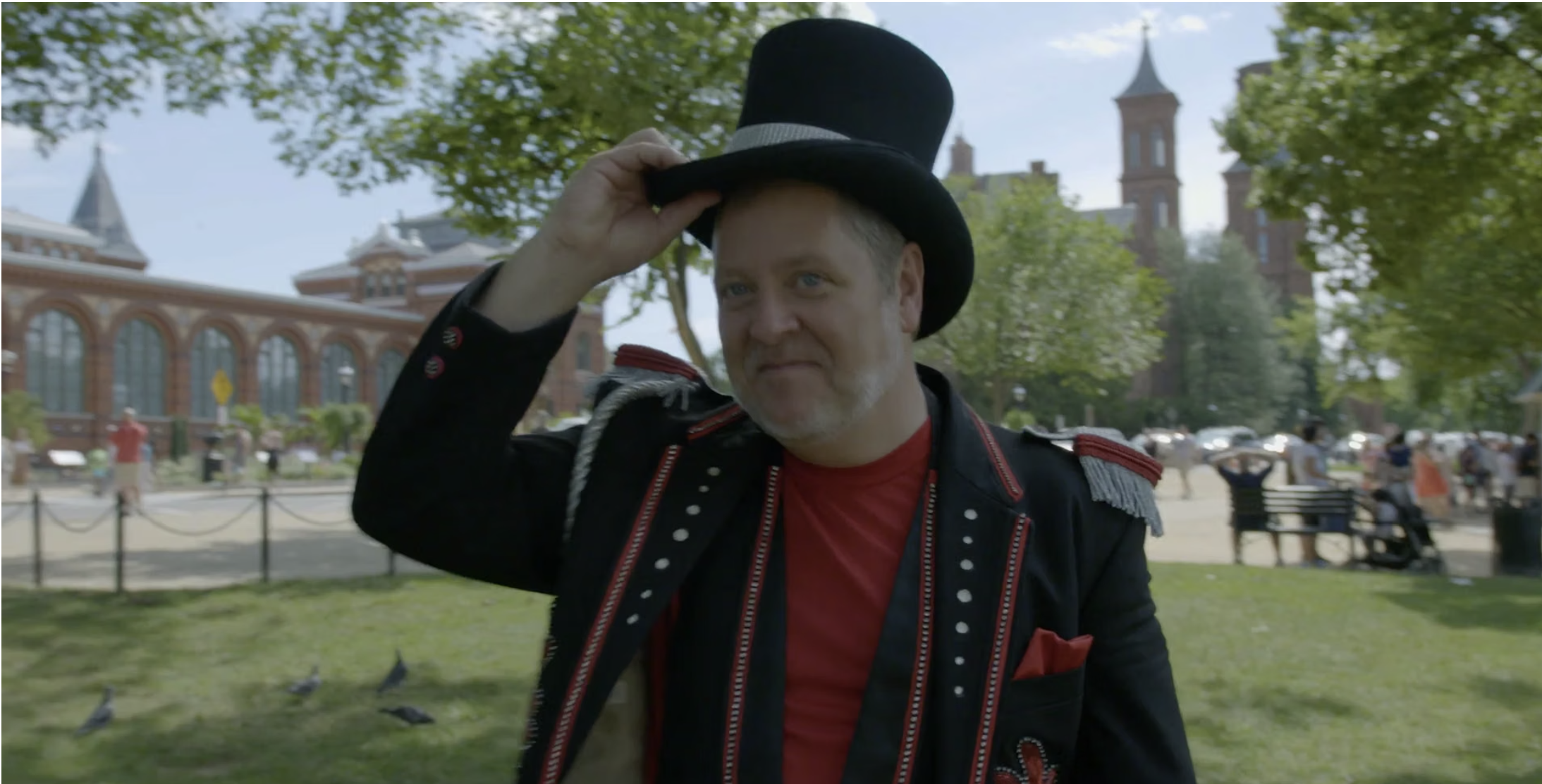 Man dressed as a circus ringmaster in a black jacket with red accents, red shirt, and a black top hat, standing outdoors in a park with people, benches, trees, and historic buildings in the background.