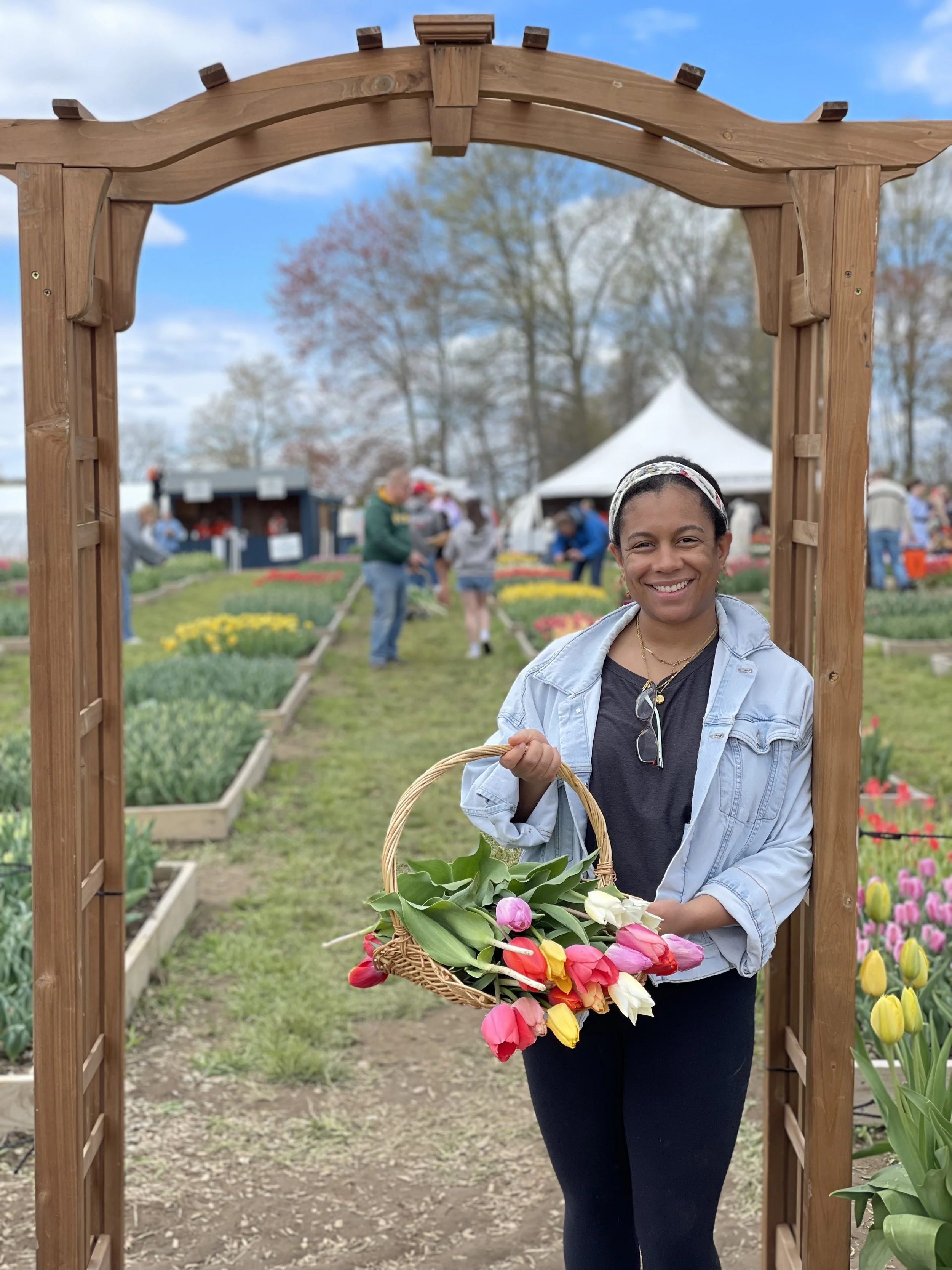 A woman smiling and holding a basket of tulips in a garden or flower festival, standing in front of a wooden arch.