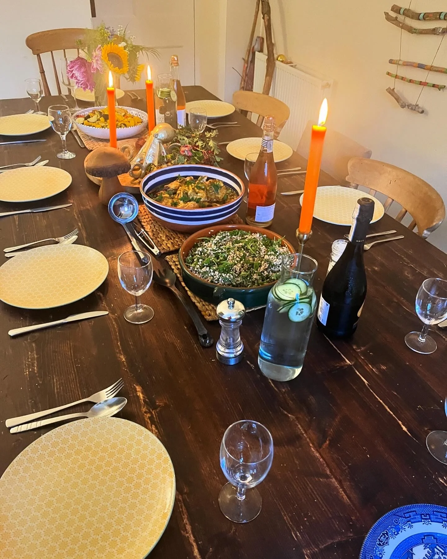 A large wooden dining table set for a meal with yellow patterned plates, silverware, empty wine glasses, and lit orange candles. There are bowls of salad and pasta, bottles of beverages, and a small vase with flowers. The background shows wooden chai
