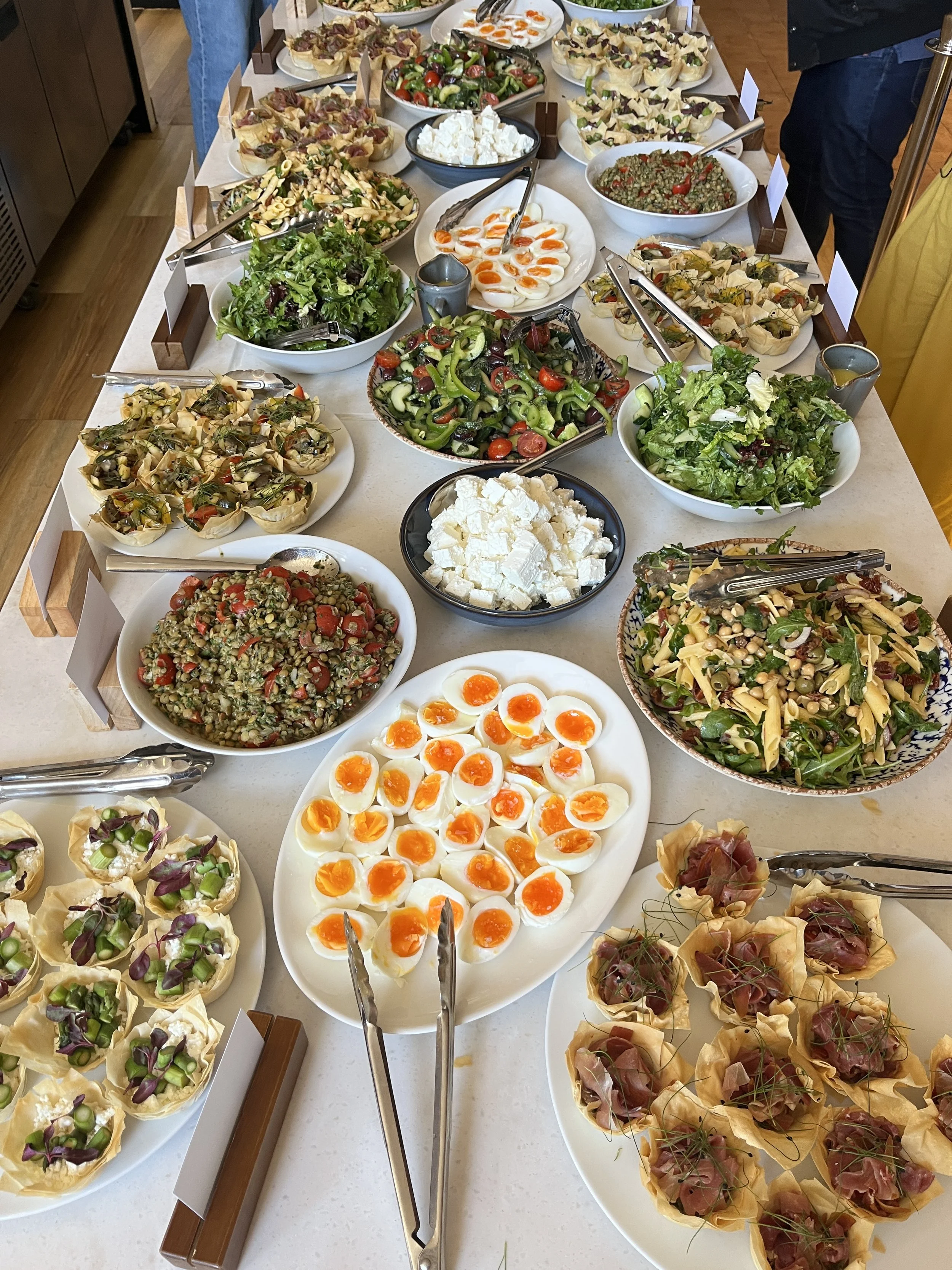A long table with a variety of salads and appetizers, including sliced hard-boiled eggs, feta cheese, mixed greens, pasta salads, and mini tartlets with various toppings. Tongs are placed next to each dish for serving.