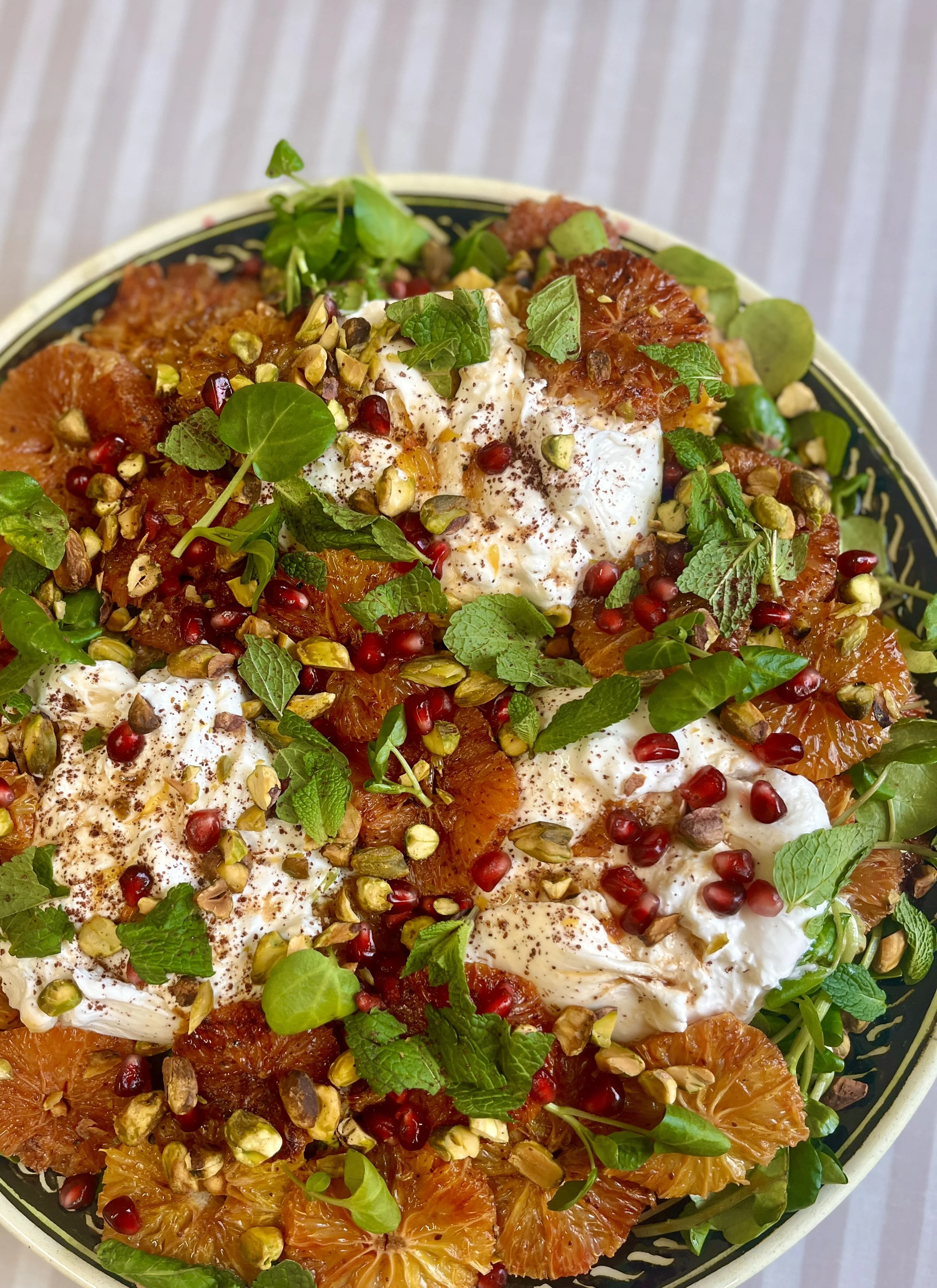 A colorful salad with orange segments, pomegranate seeds, chopped pistachios, fresh herbs, and dollops of yogurt in a bowl.