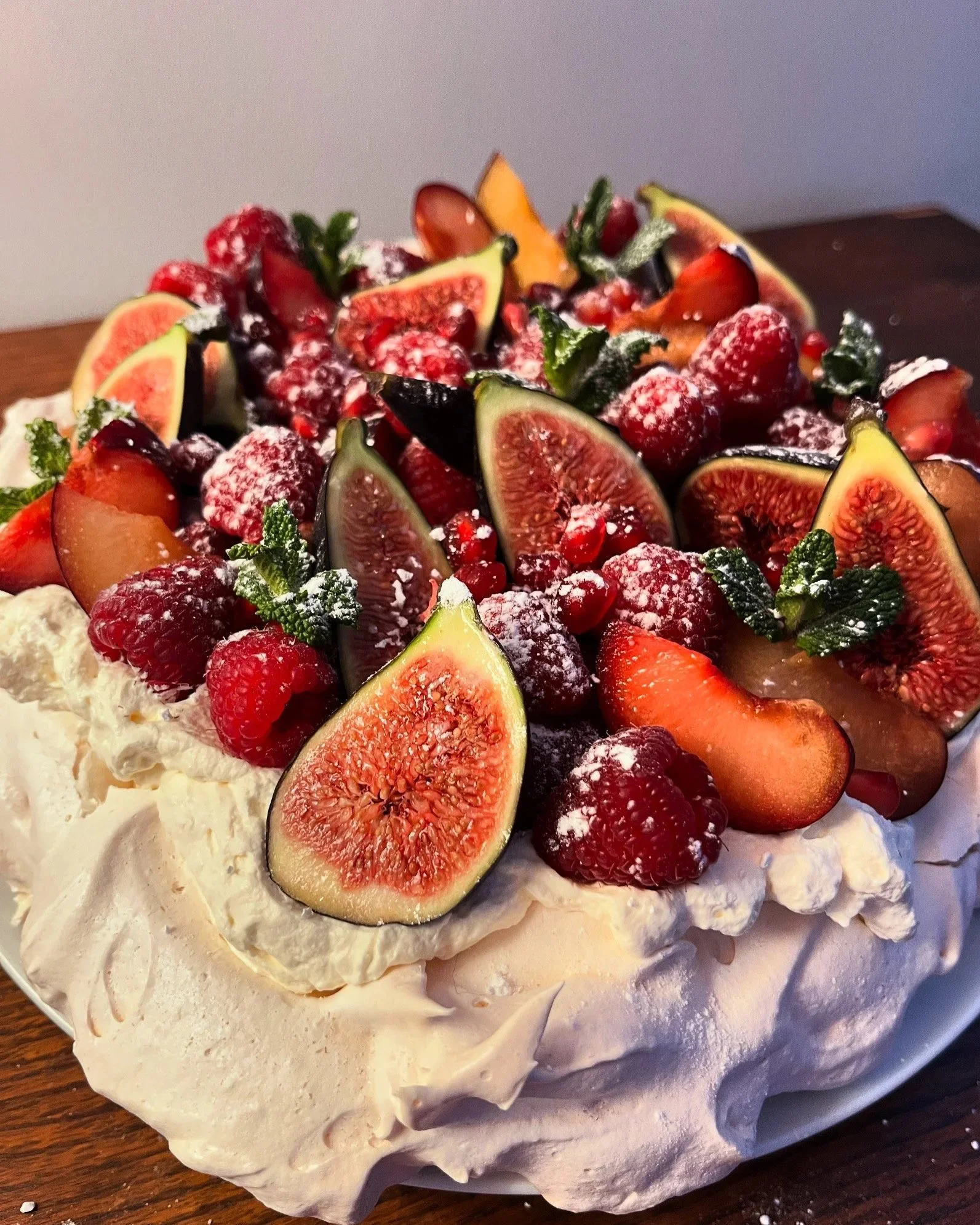 A close-up of a fruit-topped pavlova cake with sliced figs, strawberries, raspberries, pomegranate seeds, and mint leaves on a meringue base.