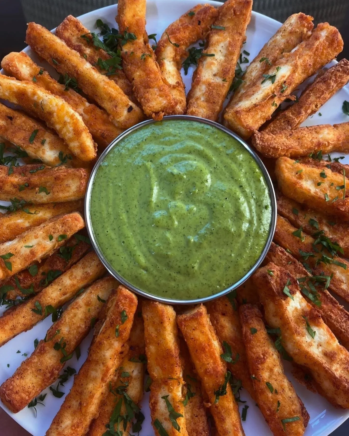 Fried mozzarella sticks garnished with chopped herbs, served with a cup of green dipping sauce in the center of a round white plate.