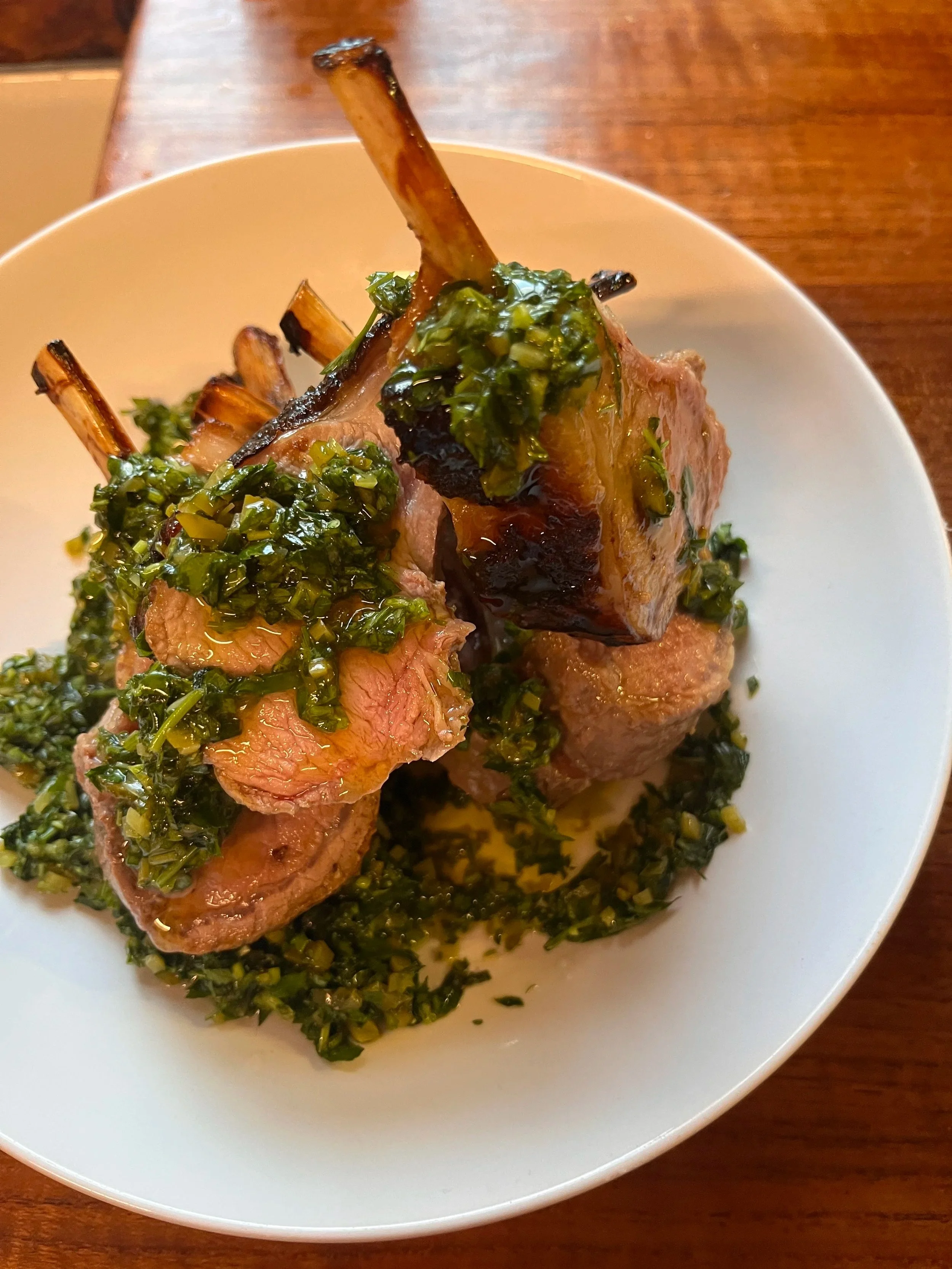 A close-up of a plated dish with two cooked lamb chops topped with green sauce and chopped herbs, surrounded by more chopped herbs on the white plate, placed on a wooden table.