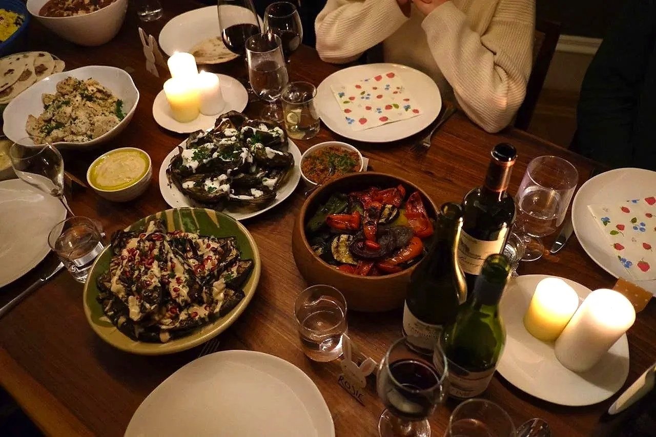 A dinner table with various dishes, including roasted vegetables, a salad, and dips, surrounded by glasses of wine and candles.