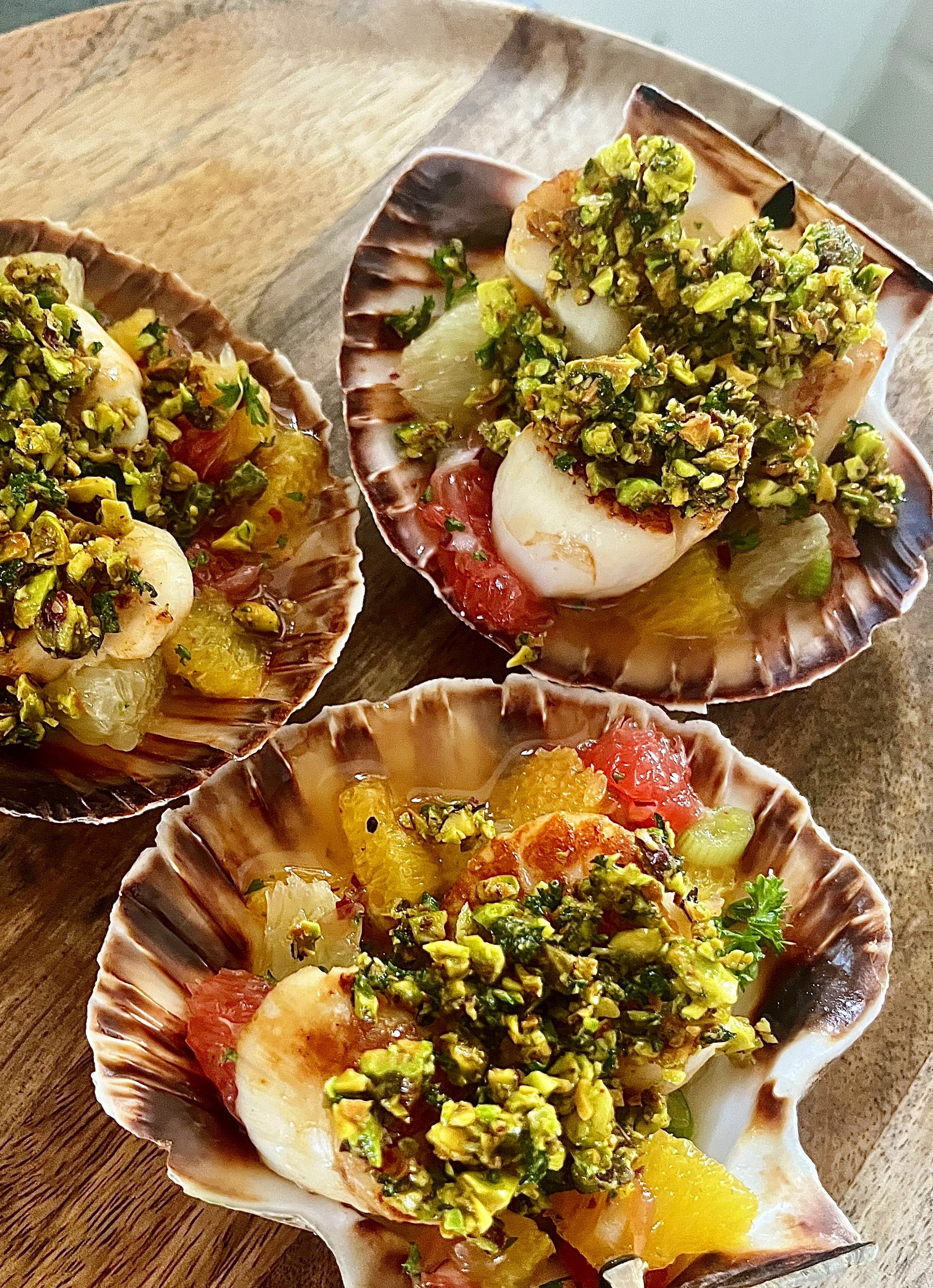 Scallop shells filled with chopped pistachios, diced tomatoes, and various chopped herbs, resting on a wooden surface.