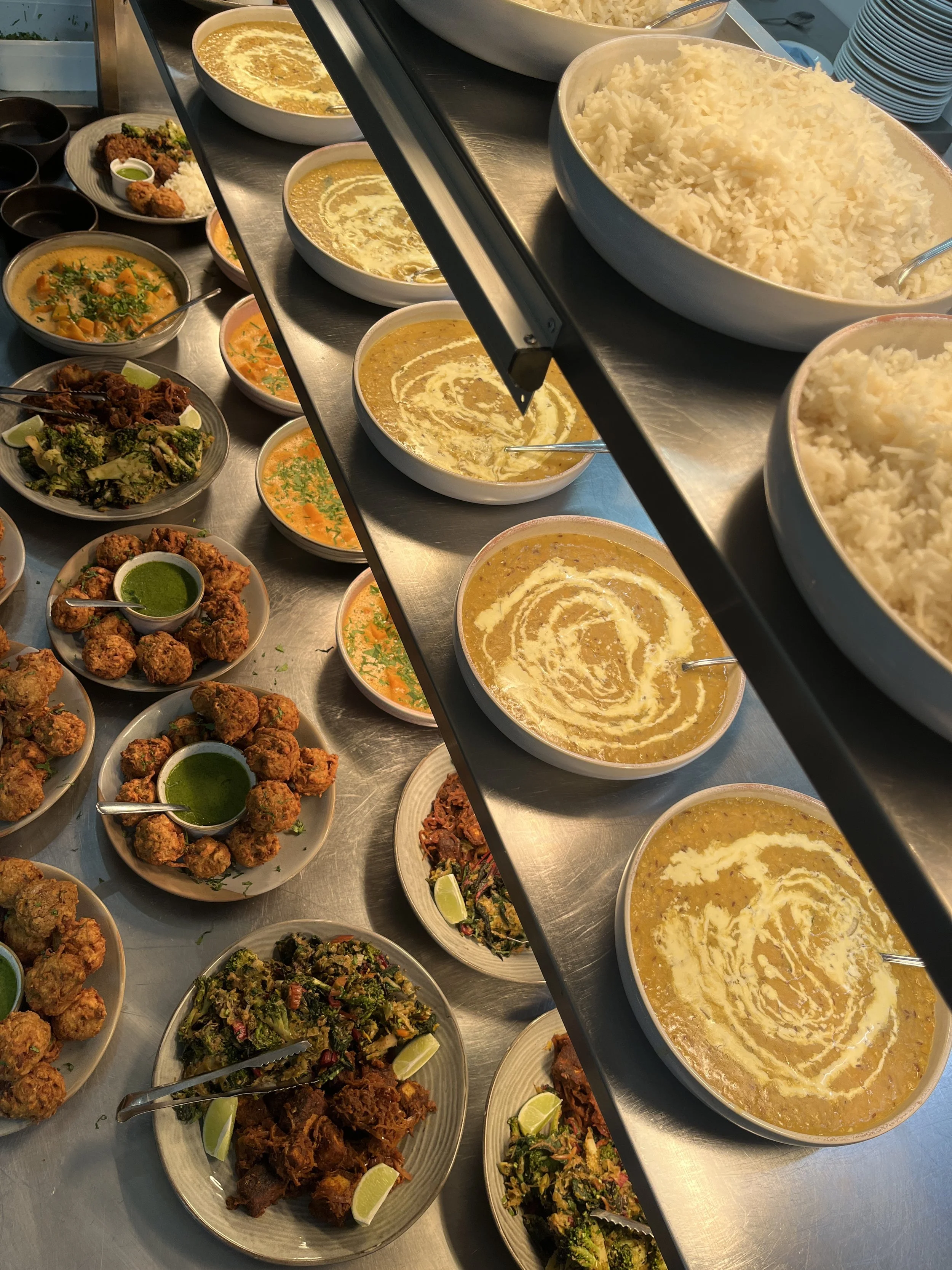 A buffet of Indian dishes including bowls of rice, curries, and fried appetizers on a metal counter.