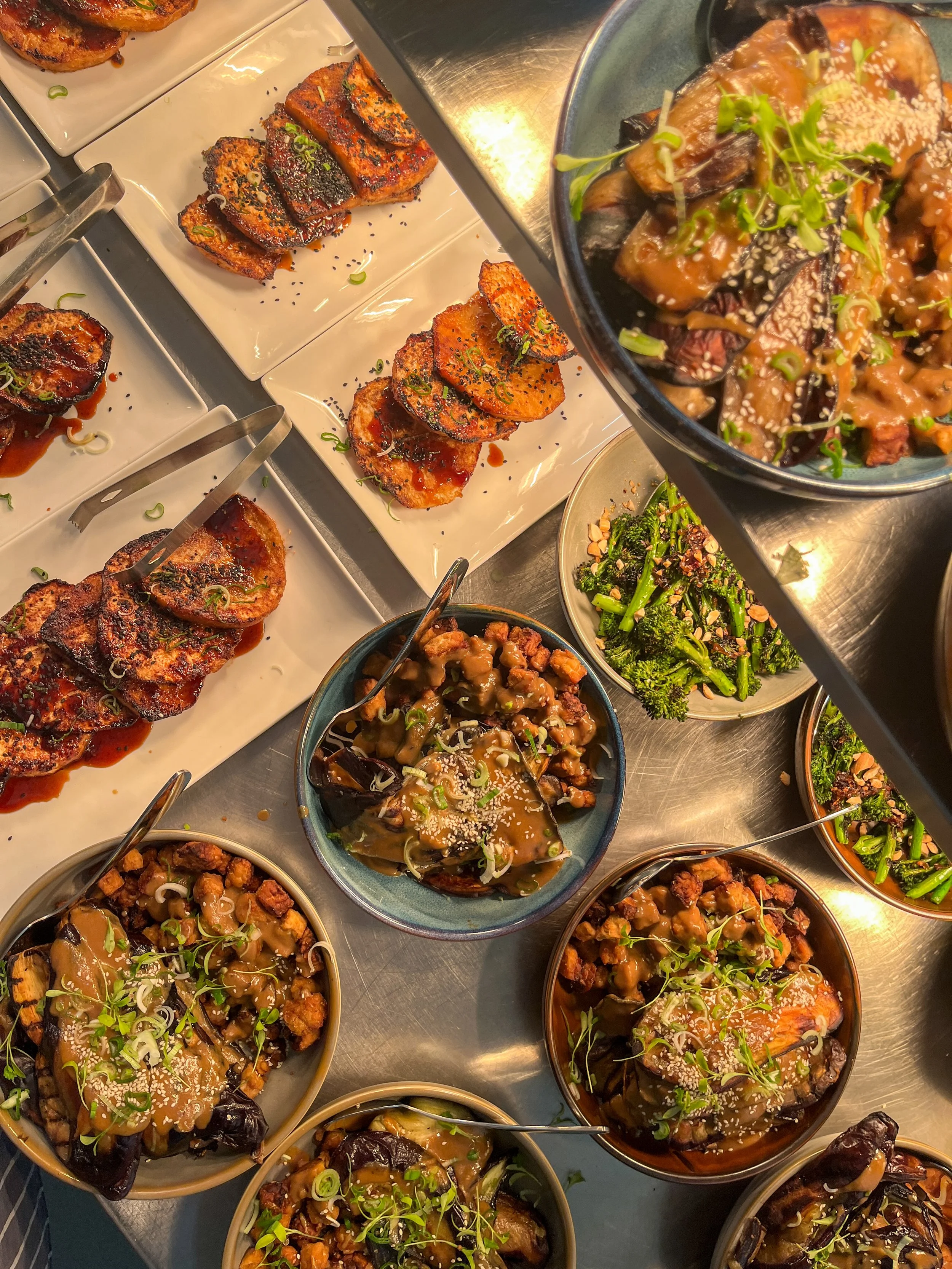 A variety of Chinese eggplant dishes topped with sesame seeds and chopped green onions, served in small bowls on a metal counter at a buffet or restaurant.