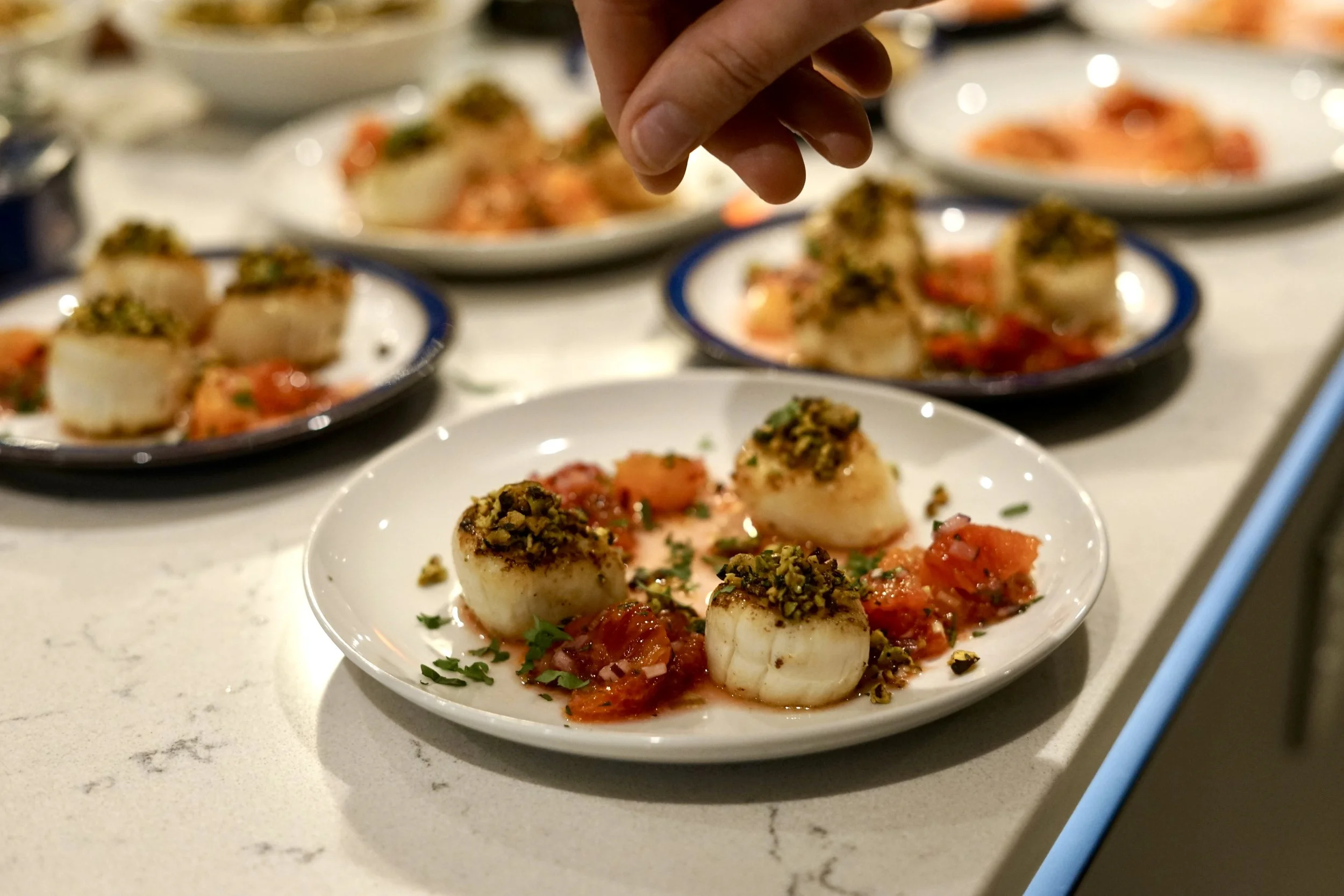 Close-up of a prepared scallop dish topped with chopped pistachios, surrounded by tomato-based sauce and garnished with herbs on a white plate.