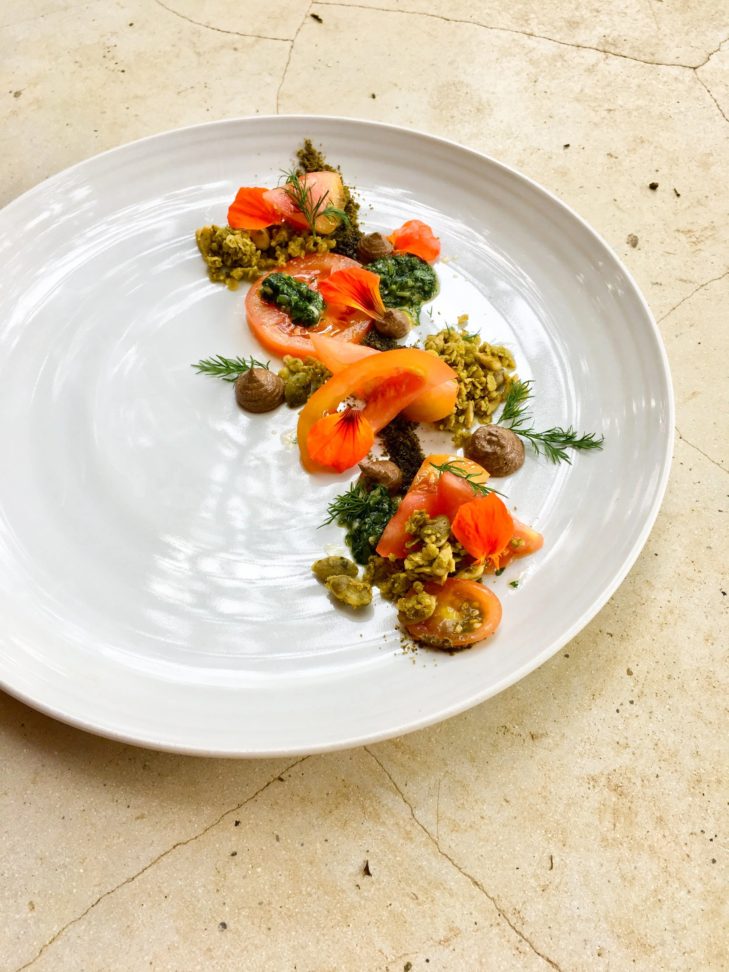 A white oval plate with a gourmet dish featuring sliced tomatoes, green sauce, edible flowers, herbs, and various garnishes, placed on a beige, cracked surface.