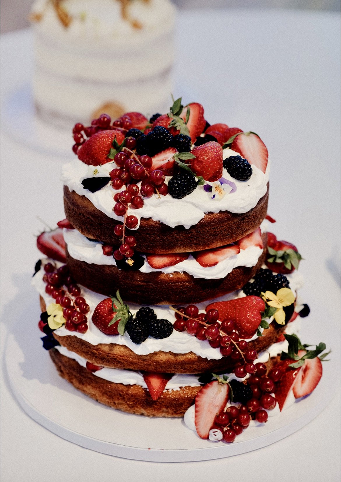 A four-tiered naked cake decorated with whipped cream and fresh strawberries, blackberries, red currants, and edible flowers.