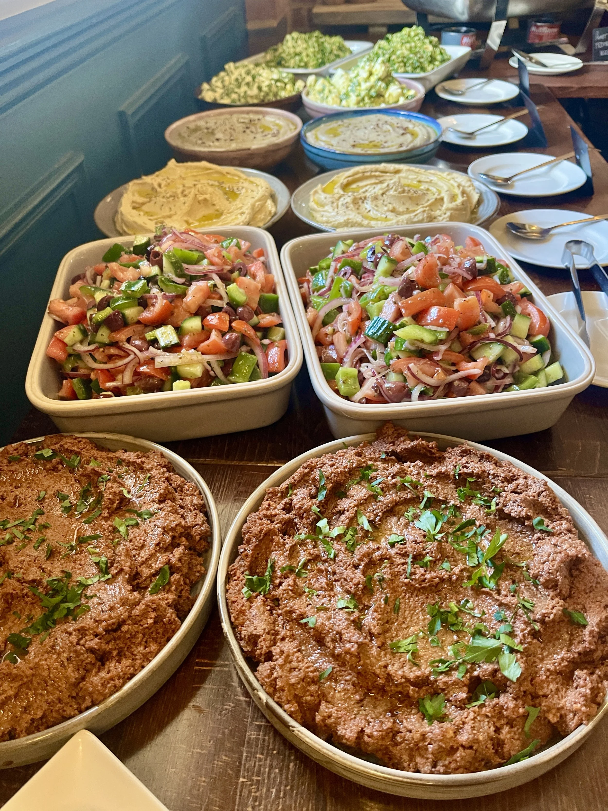 Tray of cooked meatloaf garnished with chopped parsley, accompanied by two bowls of chopped vegetable salad, and several bowls of dips and side dishes, on a wooden table in a restaurant setting.