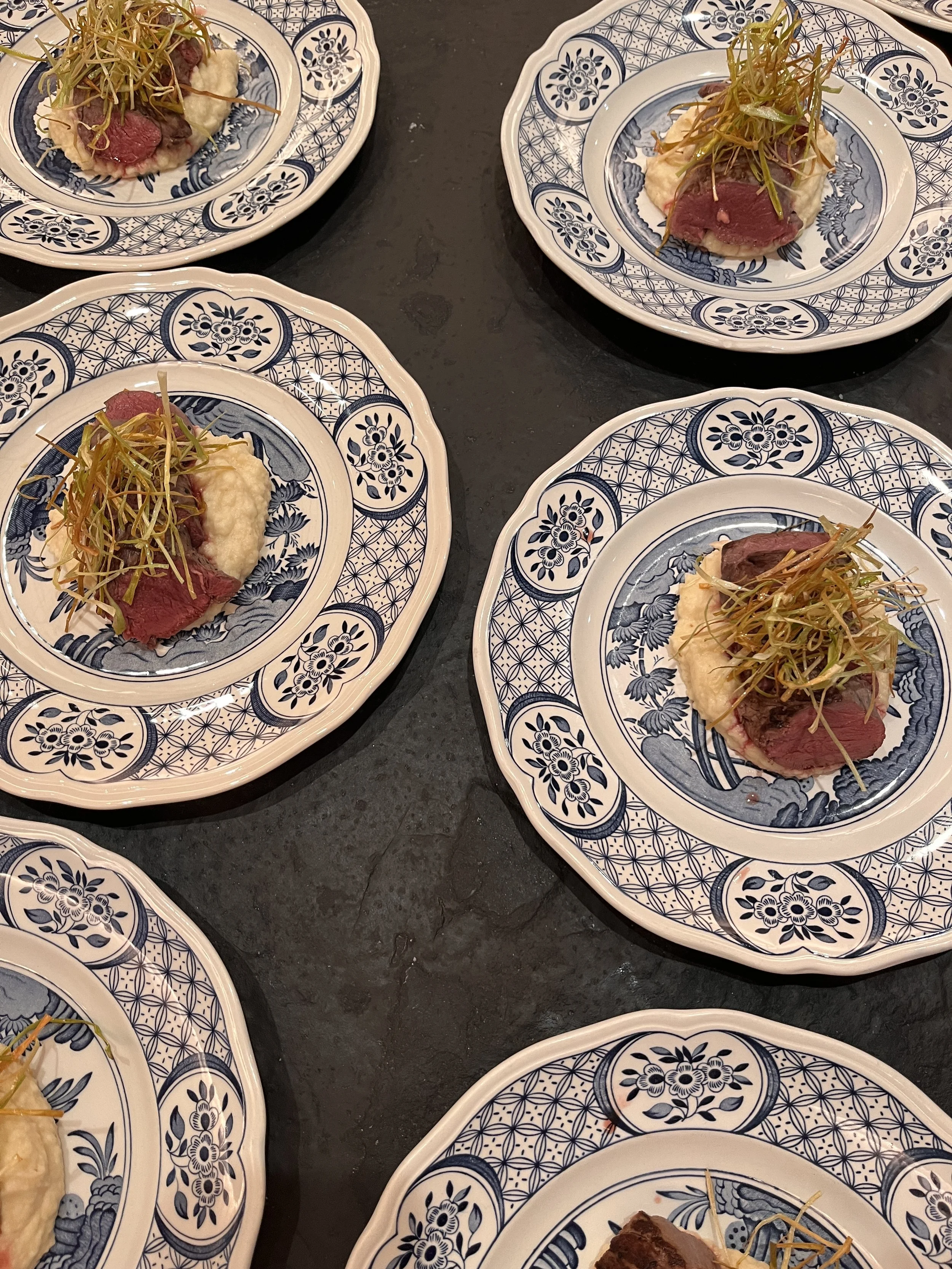 Multiple blue and white patterned plates with slices of cooked red meat, mashed potatoes, and garnished with microgreens, placed on a black table.