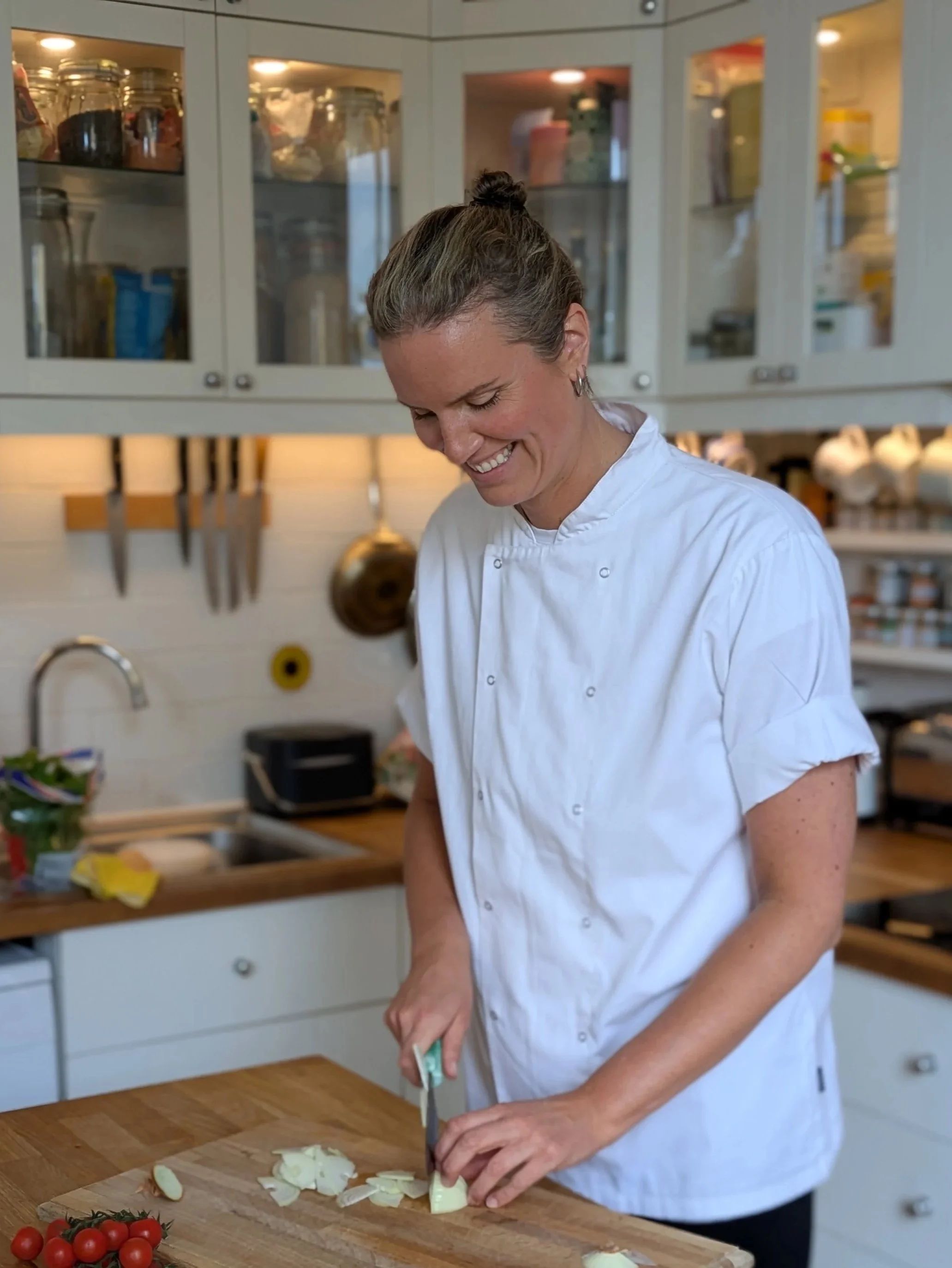 A woman private chef is chopping onions on a wooden cutting board in a kitchen, with cherry tomatoes on the counter and kitchen cabinets in the background.