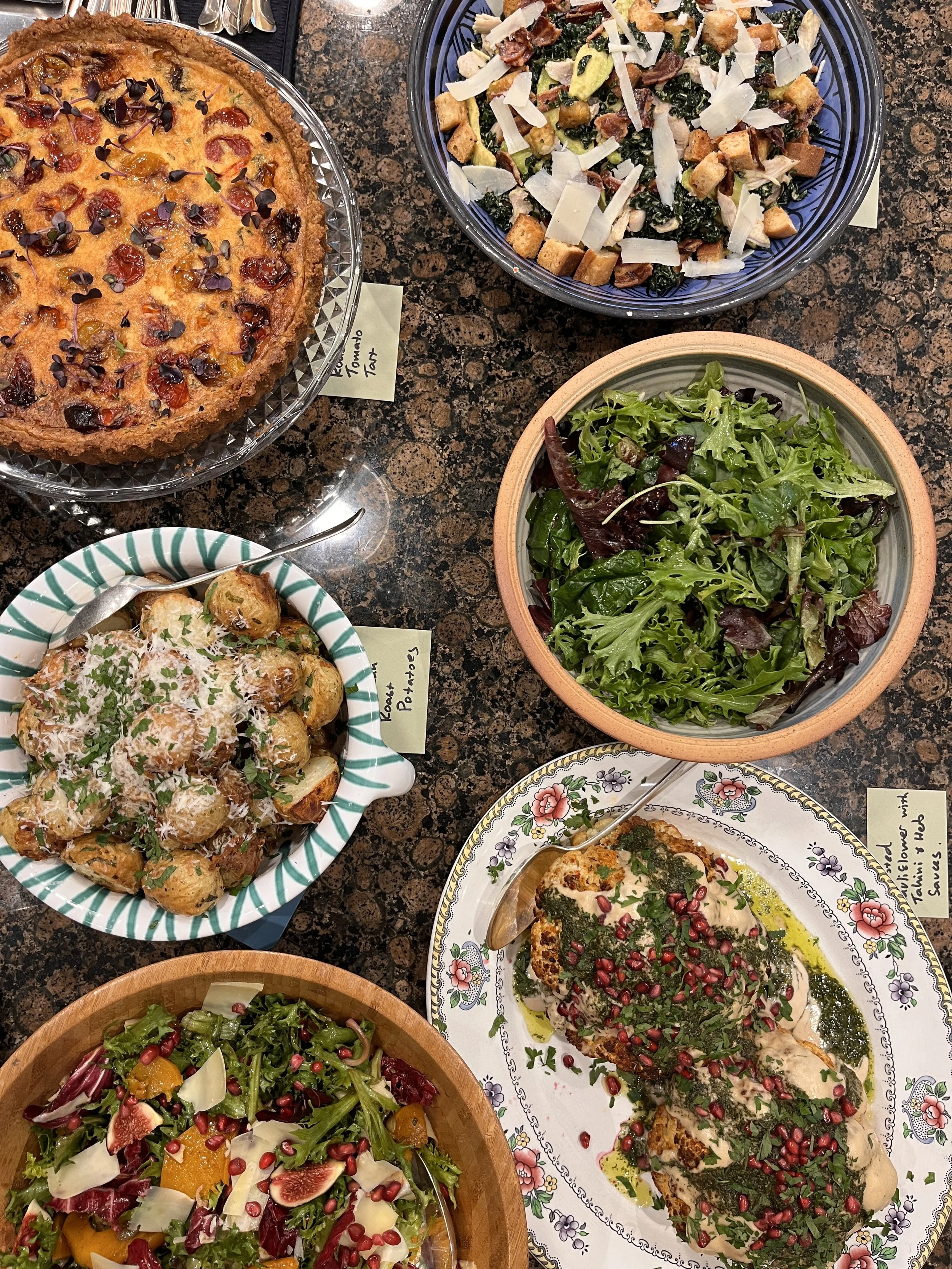 A variety of dishes on a table, including a tomato tart, potato salad, mixed green salad, roasted chicken with herbs and pomegranate seeds, and a fruit salad with figs, pomegranate seeds, and greens.